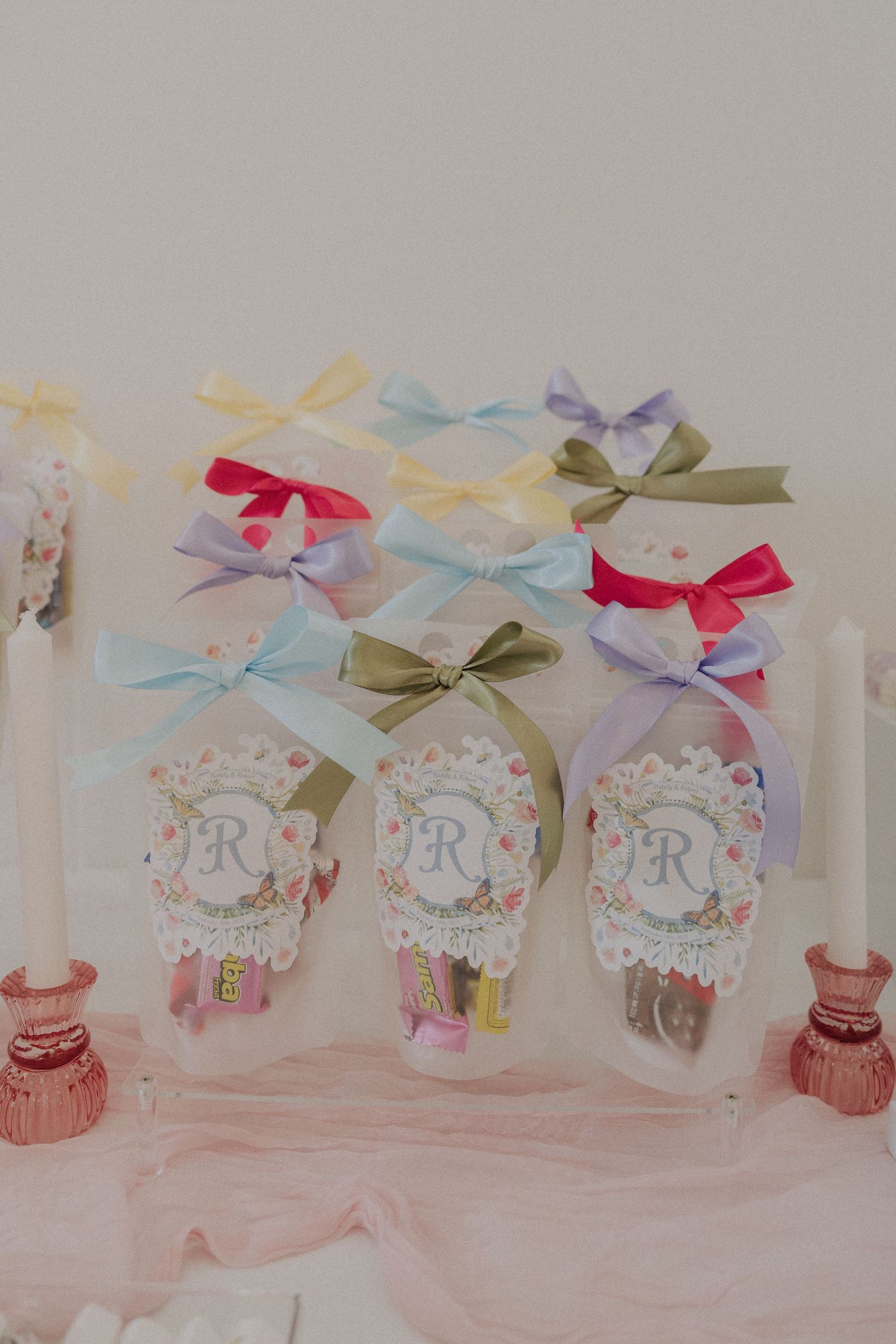 Elegant party favors with colorful ribbon bows and monogram R tags displayed on a white tray with pink candlesticks.