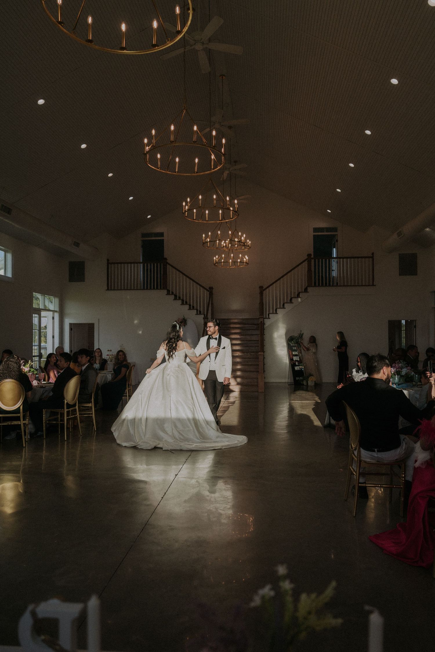 Bride and groom share their first dance in an elegant wedding venue with chandeliers and guests seated nearby.
