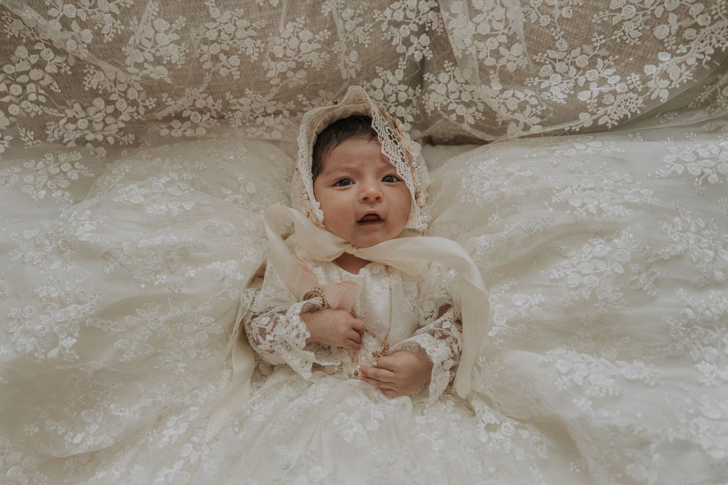 Baby wearing an ornate white christening gown lying on lace fabric.