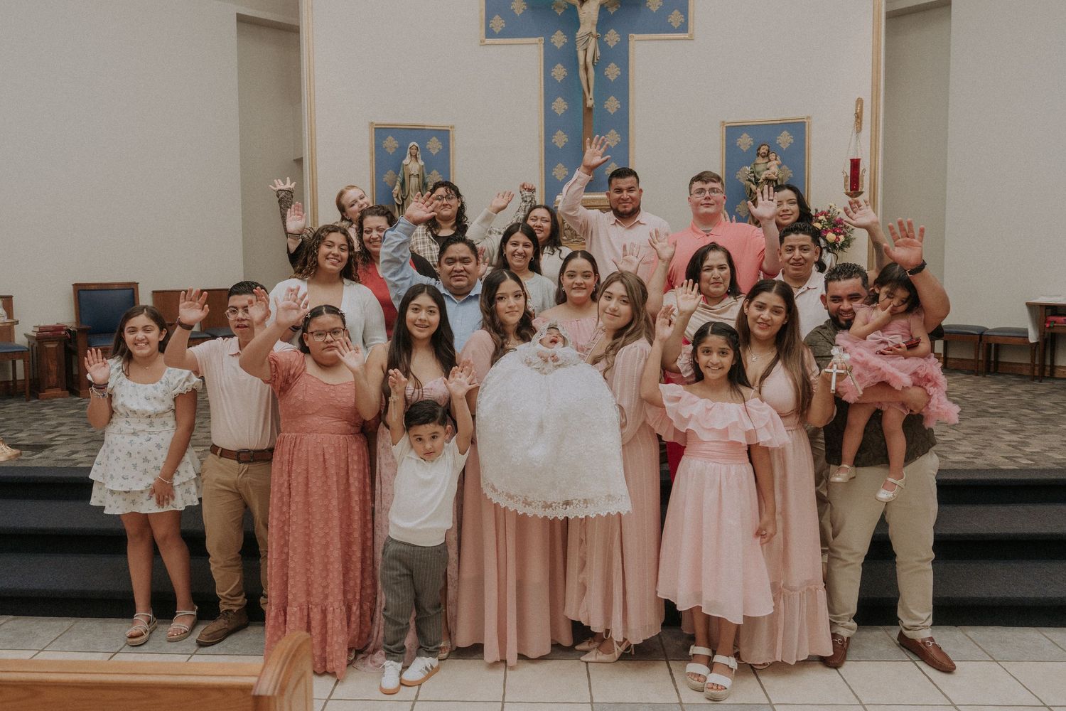 Large group photo of baptism party members wearing pink and beige attire in a church setting.