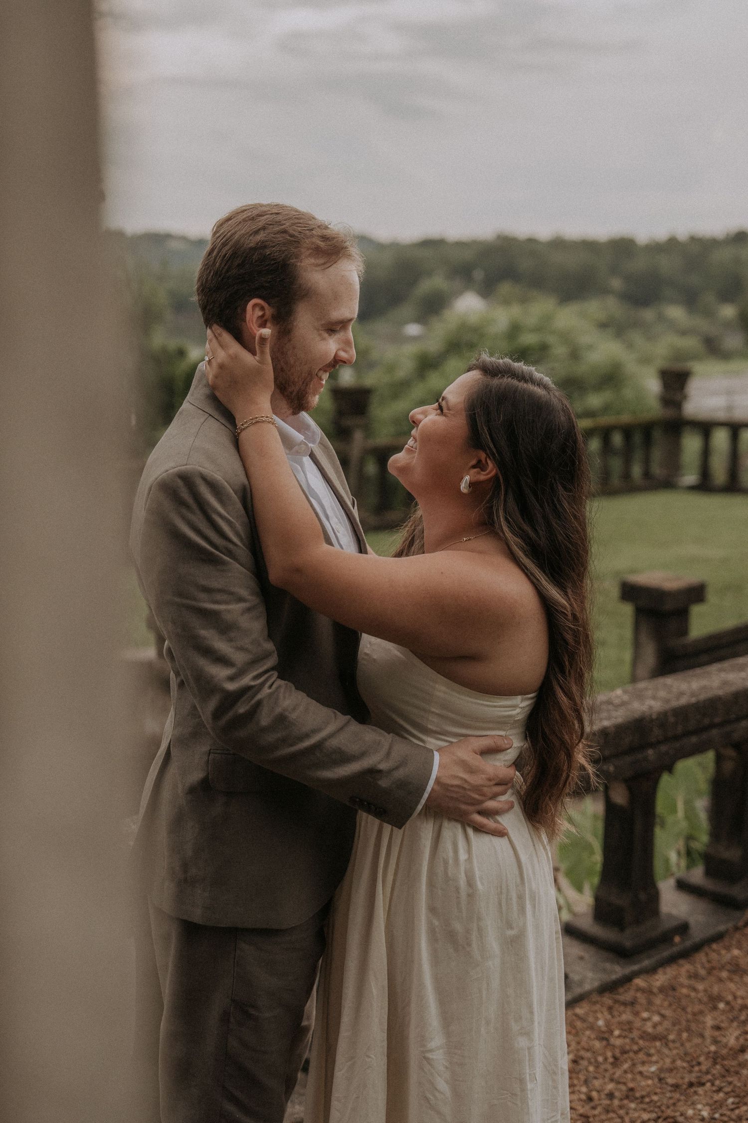 A couple shares romantic moments along a rustic stone railing surrounded by lush greenery and trees.