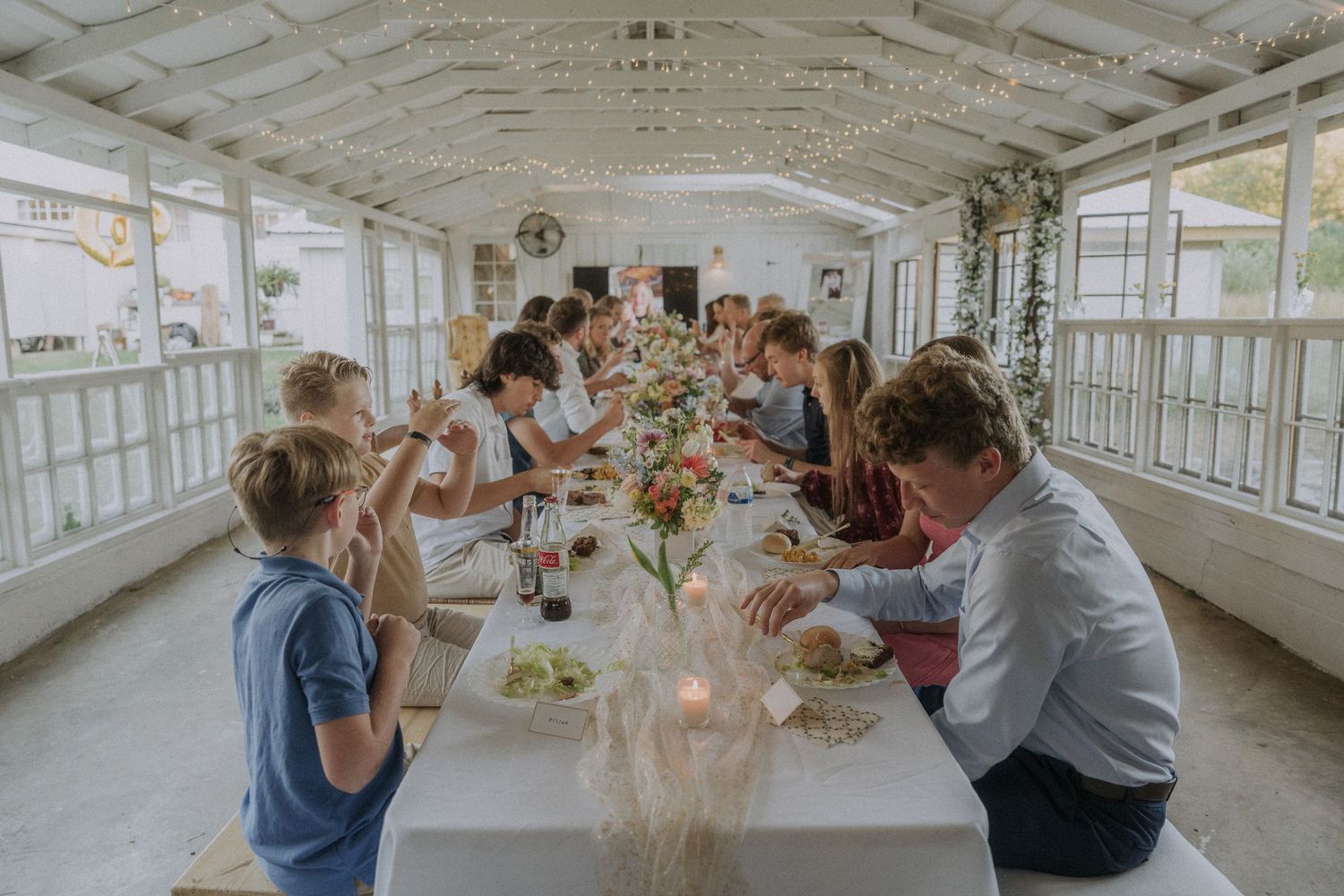 Long outdoor room with white walls and windows filled with celebration guests.
