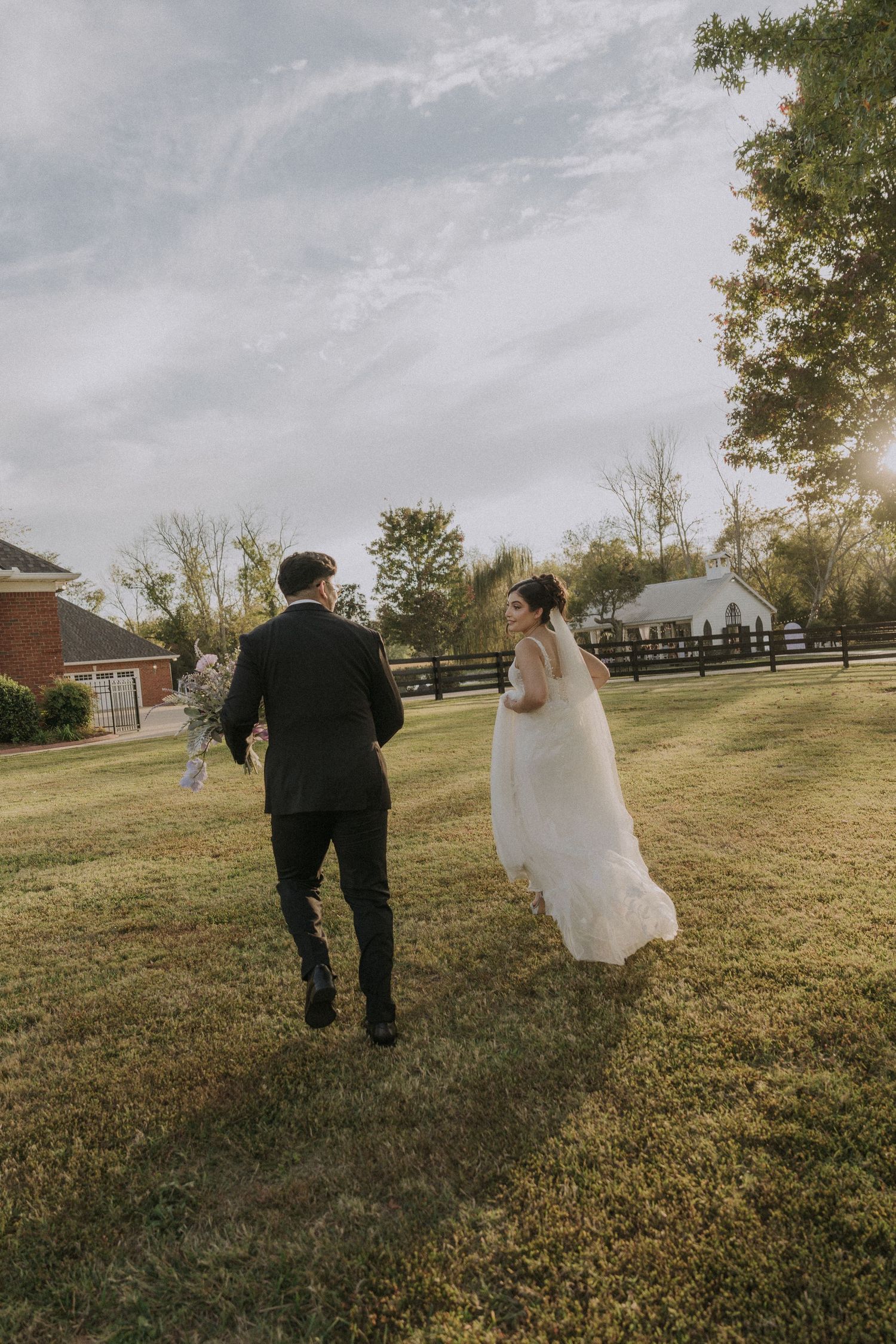 Bride and groom running hand in hand across a lush green lawn at a rustic outdoor wedding venue.