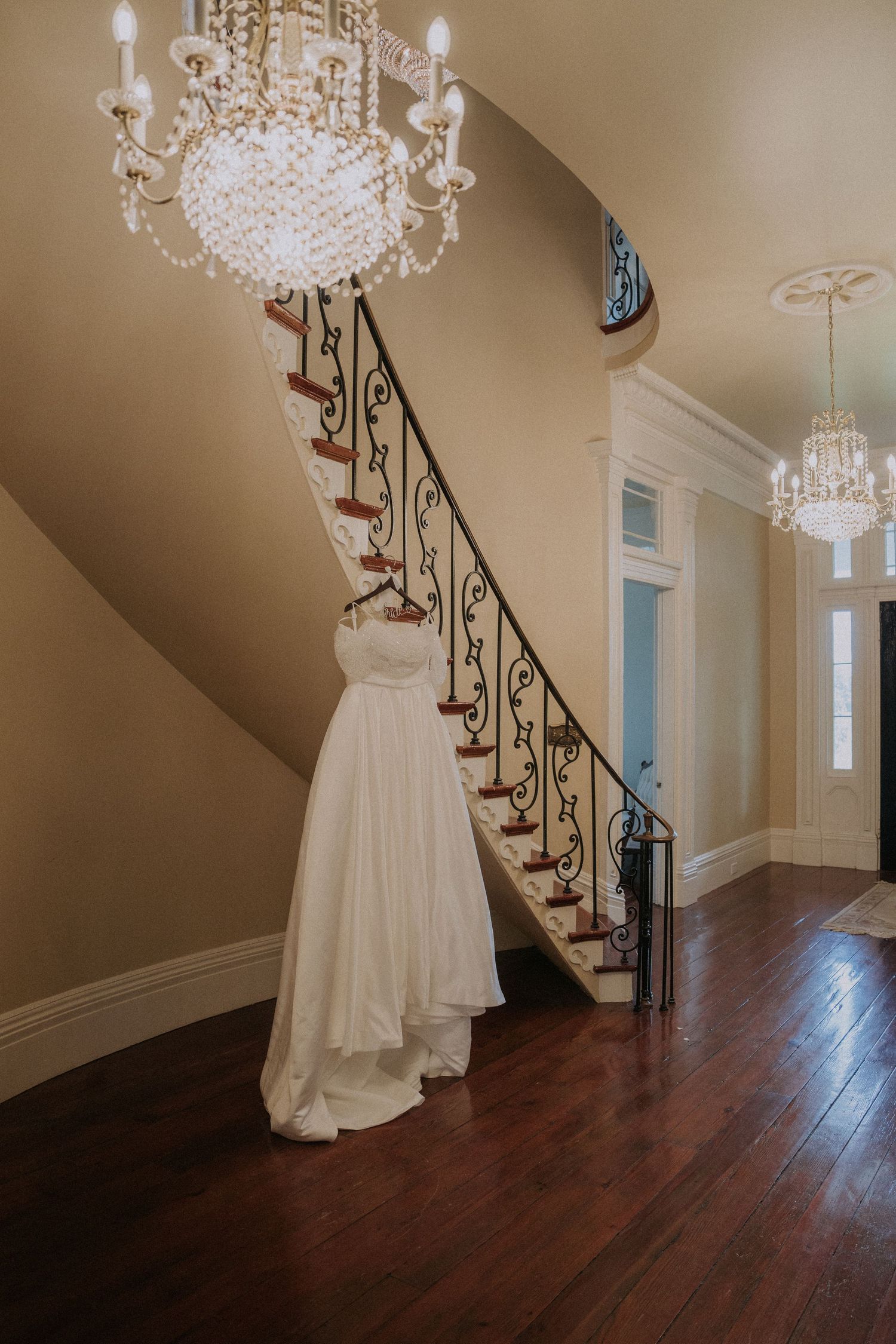 White wedding dress hanging on ornate staircase railing beneath crystal chandelier in elegant foyer.
