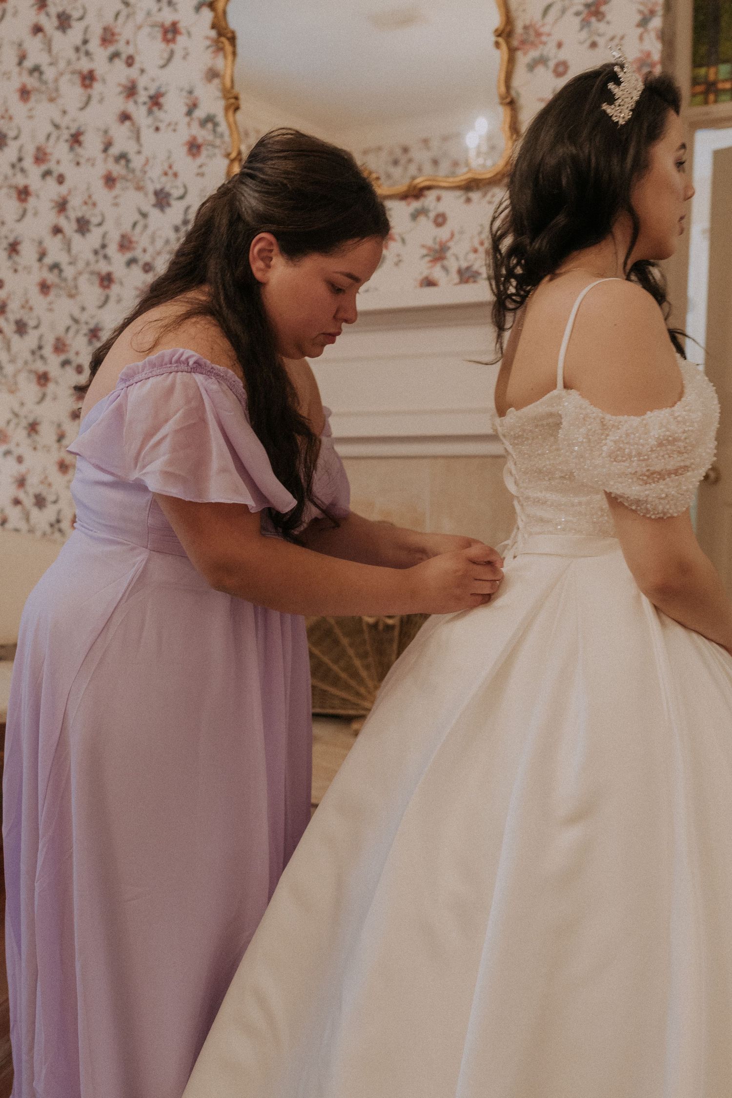Bridesmaid in lavender dress helping bride with white lace wedding gown in floral decorated dressing room.