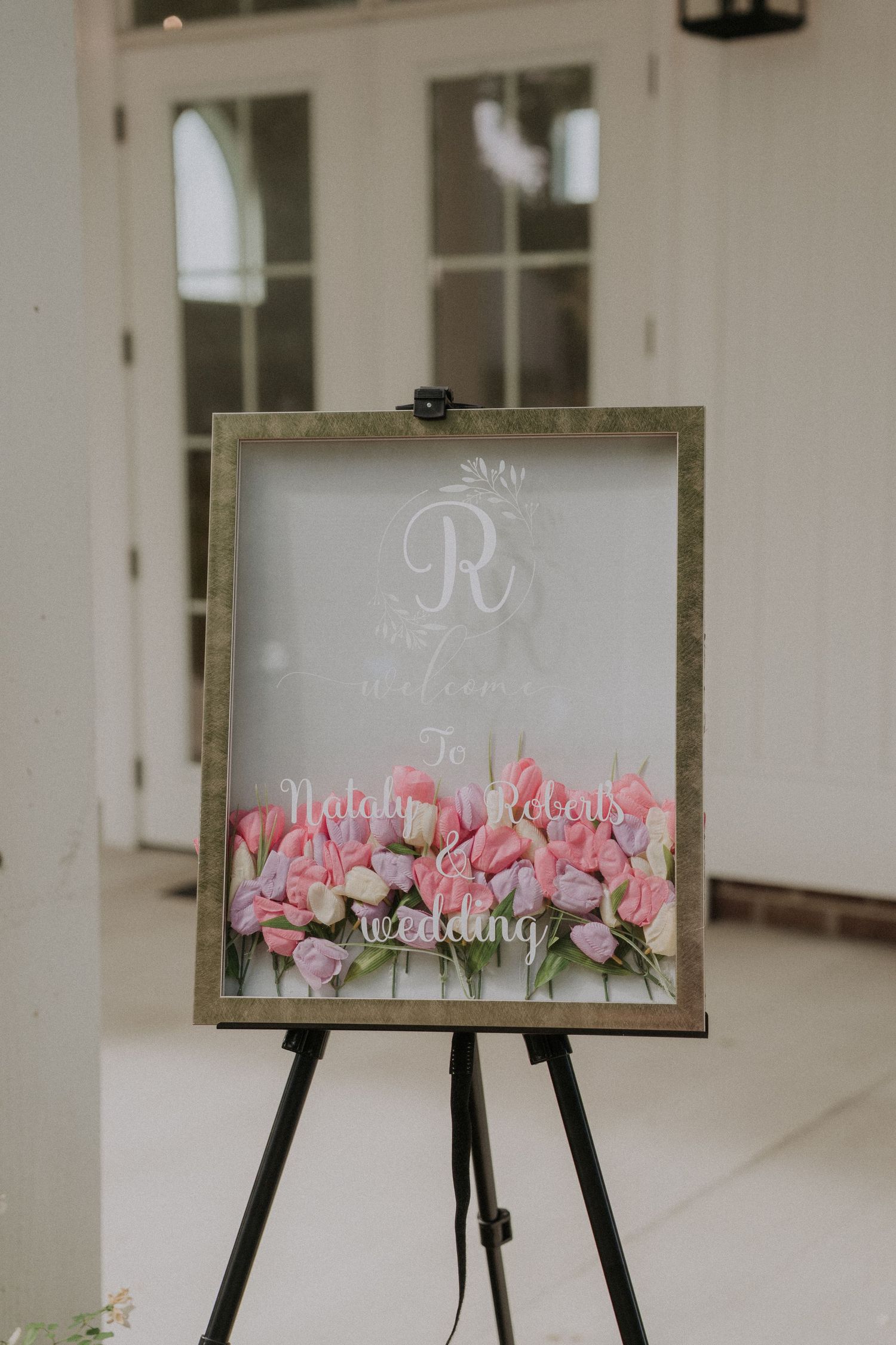 Elegant wedding welcome sign on easel with pink tulips and monogram R, displayed at venue entrance.
