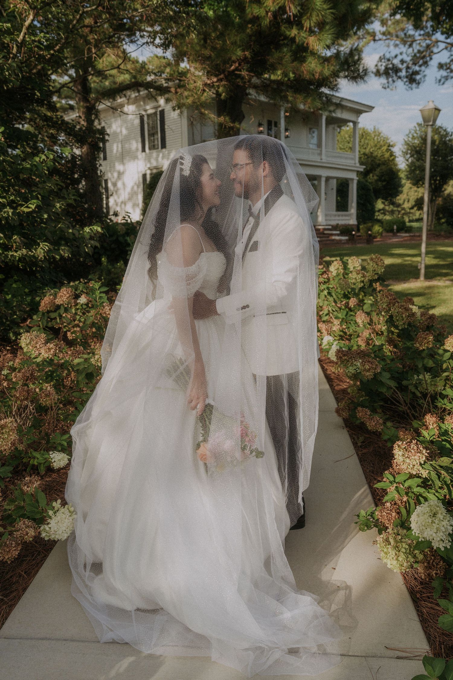 Bride and groom share a romantic kiss under a flowing veil on a garden path outside a historic white home.