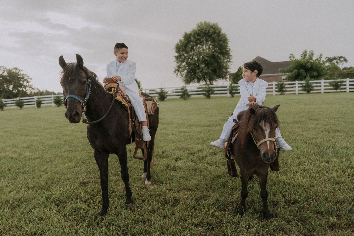Two boys in baptism clothing on horseback pose together in a scenic pasture with white fencing.