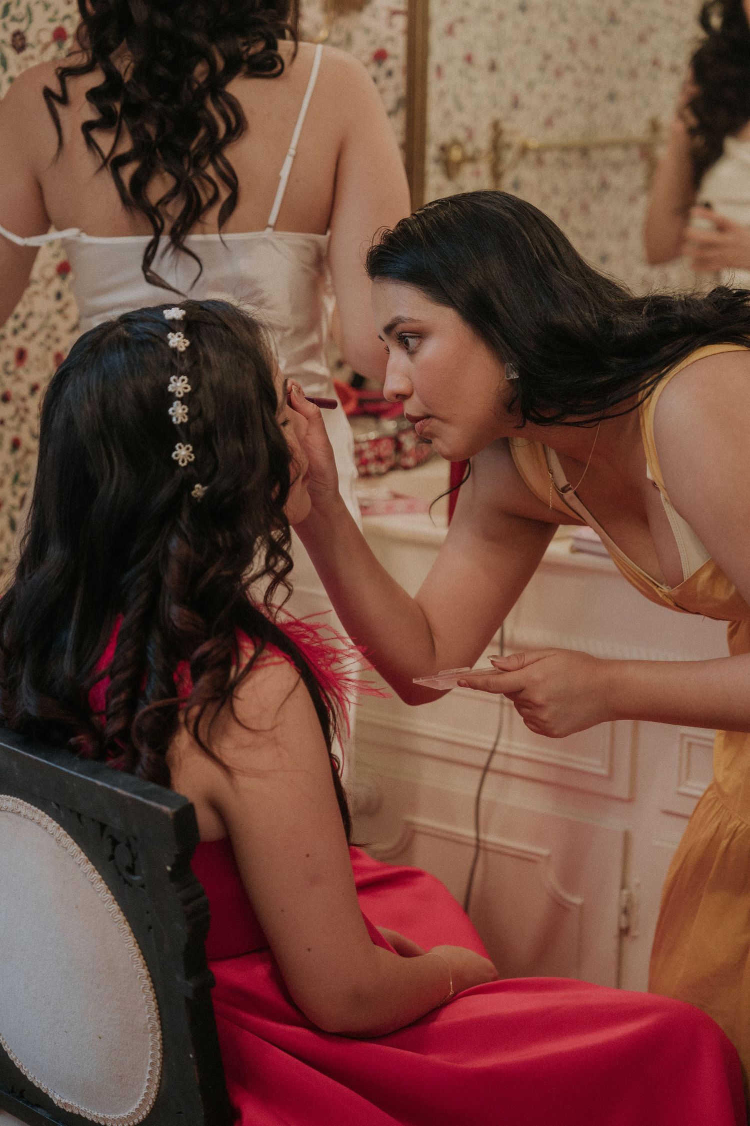 A makeup artist in a yellow dress applies makeup to a woman in a pink gown with a floral hair accessory.