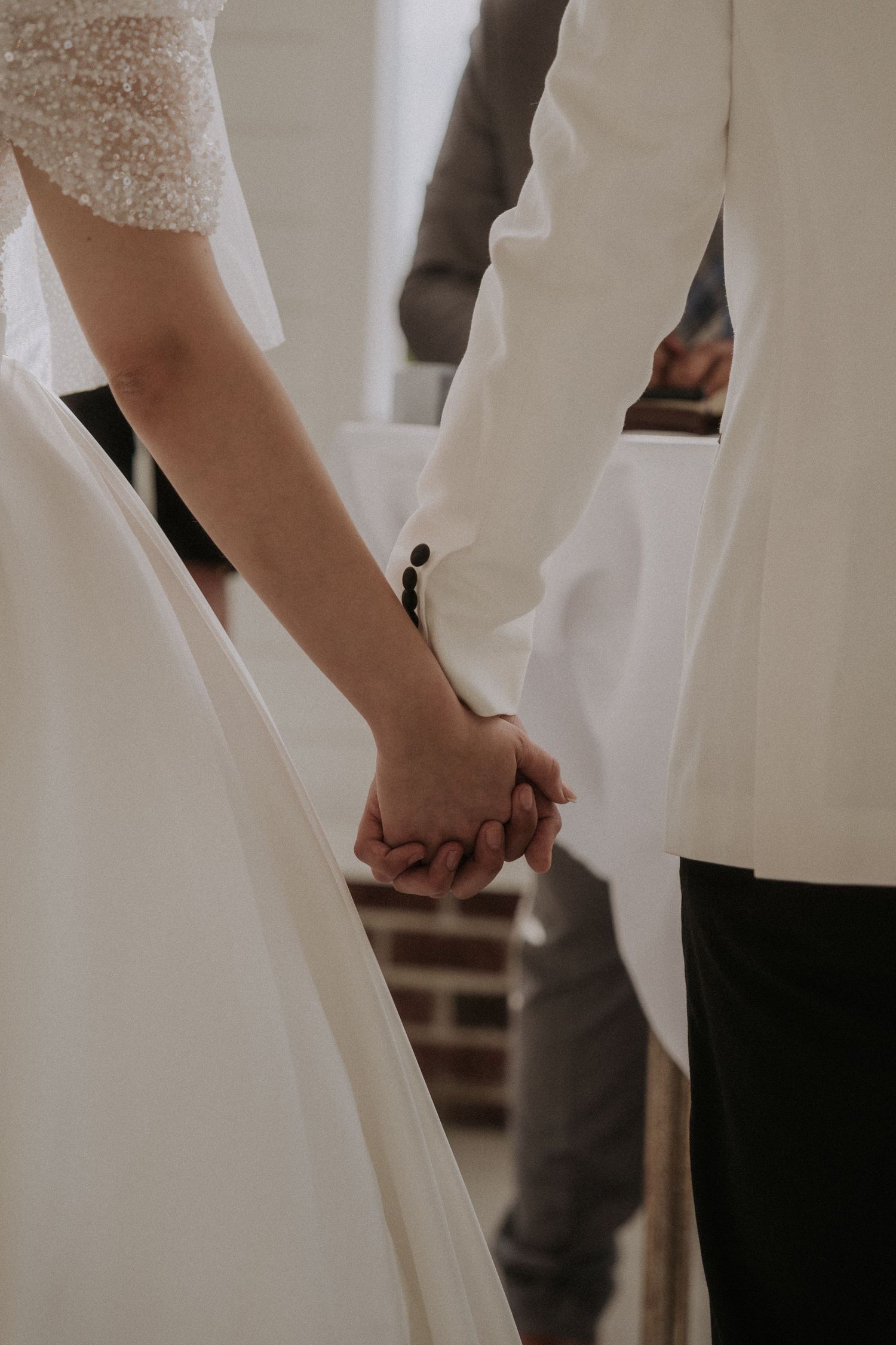 Bride and groom holding hands during wedding ceremony, bride in white lace gown, groom in white suit jacket.