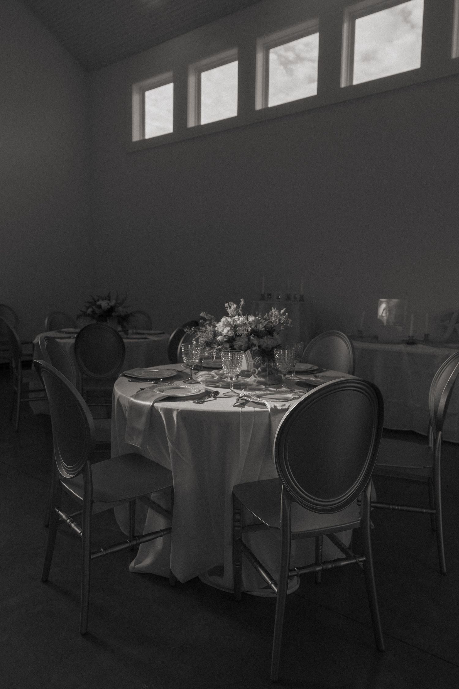 Black and white photo of an elegant round dining table set with floral centerpiece and medallion chairs in a banquet hall.