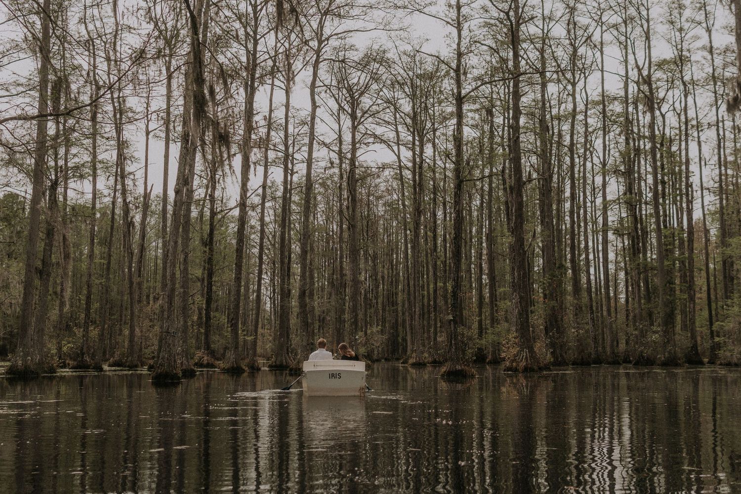 Moody landscape photograph of couple in a canoe amongst tall cypress trees reflected in dark swamp waters.