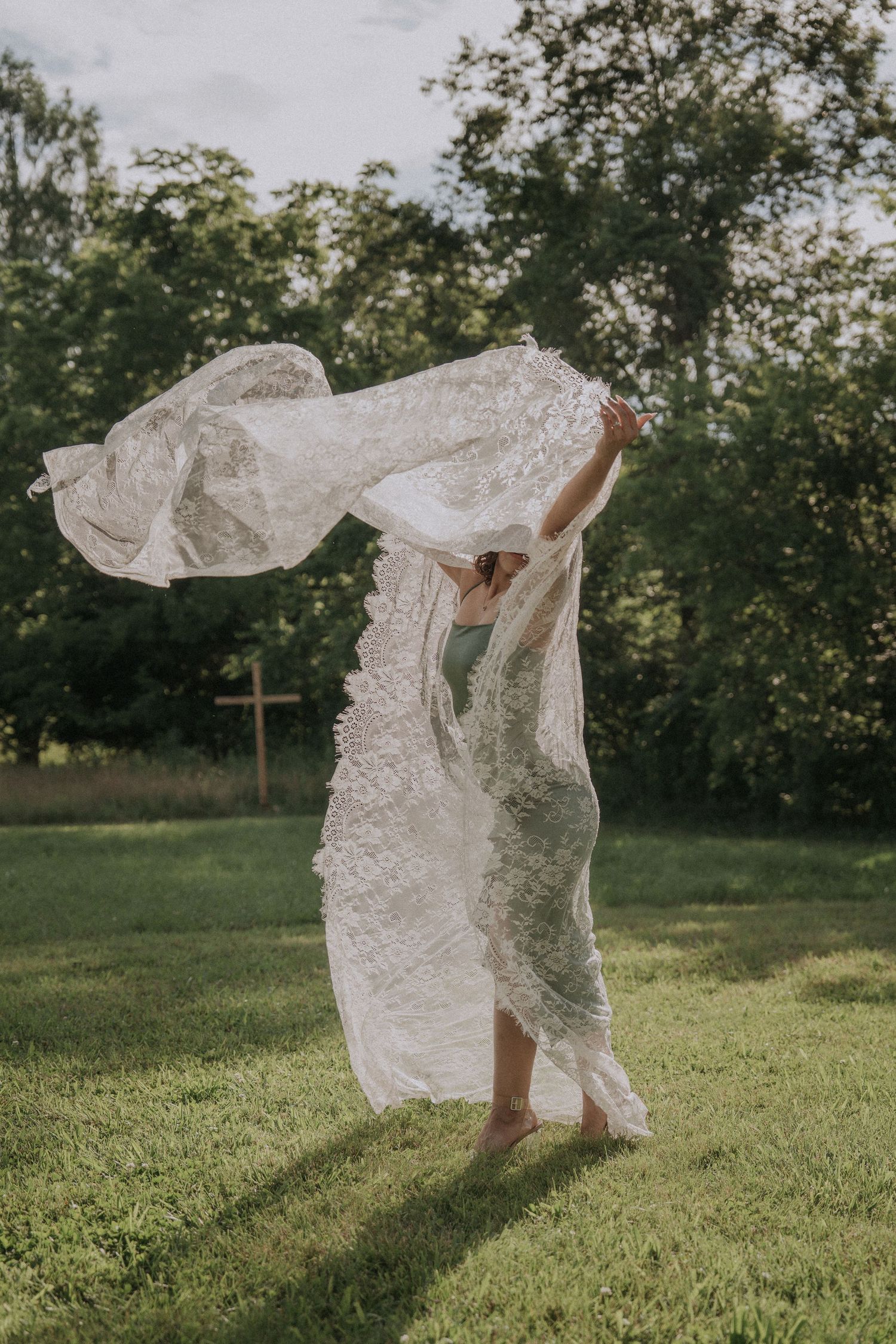 A woman standing under flowing white lace billowing in the wind against a green grassy background.