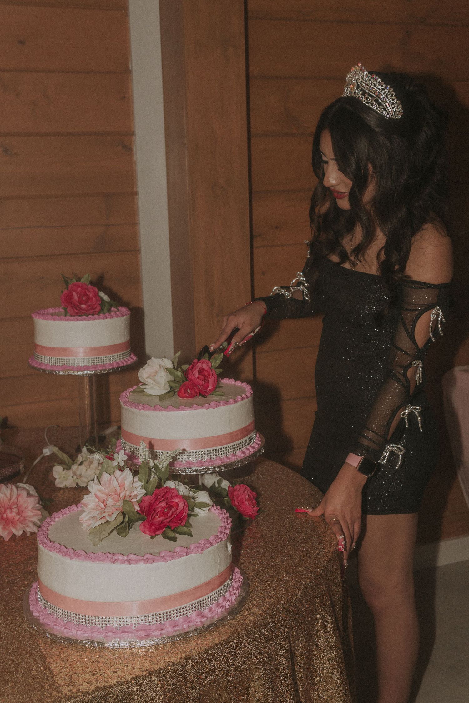 An elegant tiered white and pink cake decorated with roses sits on a gold sequined tablecloth.