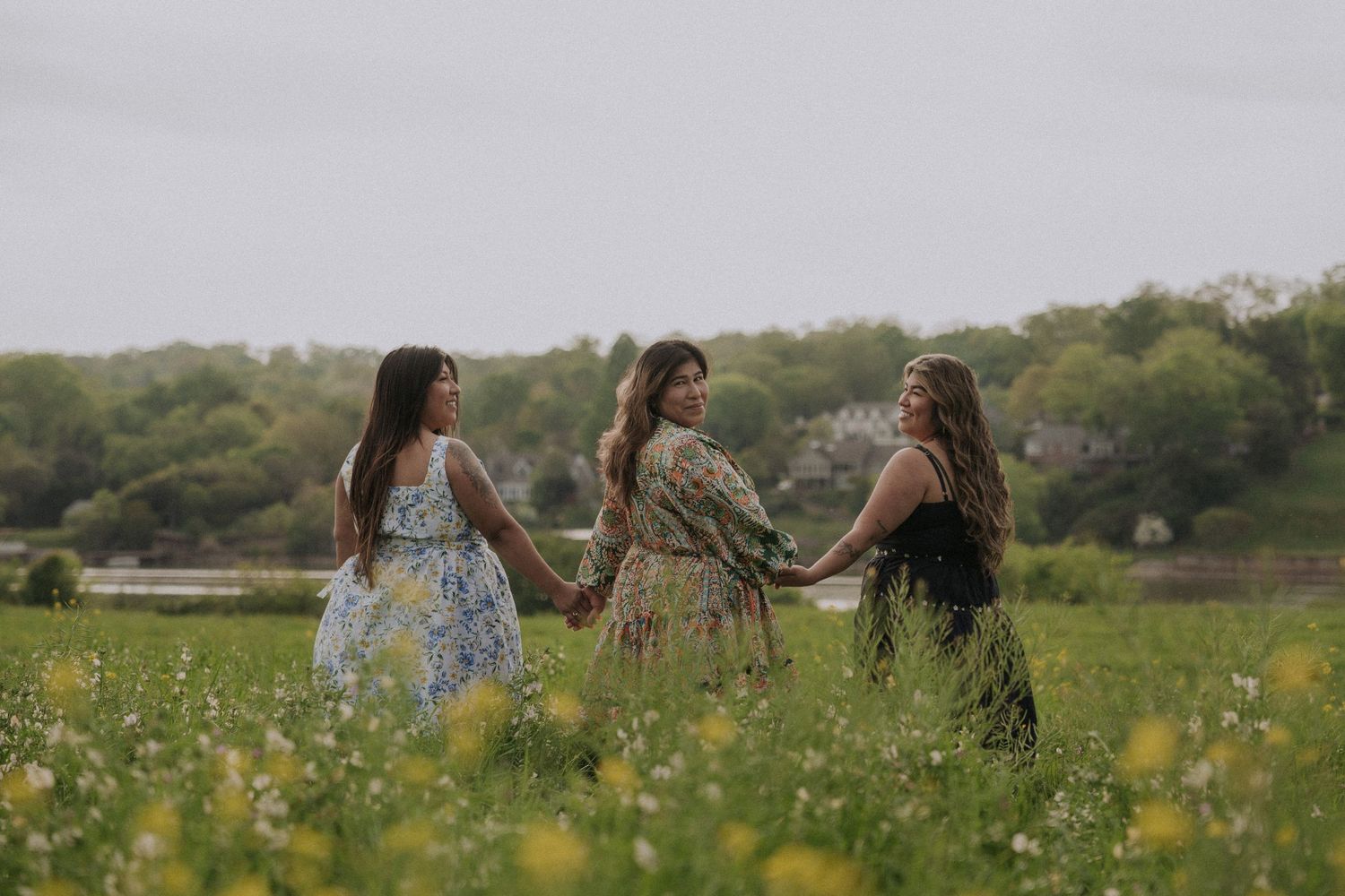 Three women in dresses hold hands in a blooming wildflower meadow, posing for an outdoor portrait.