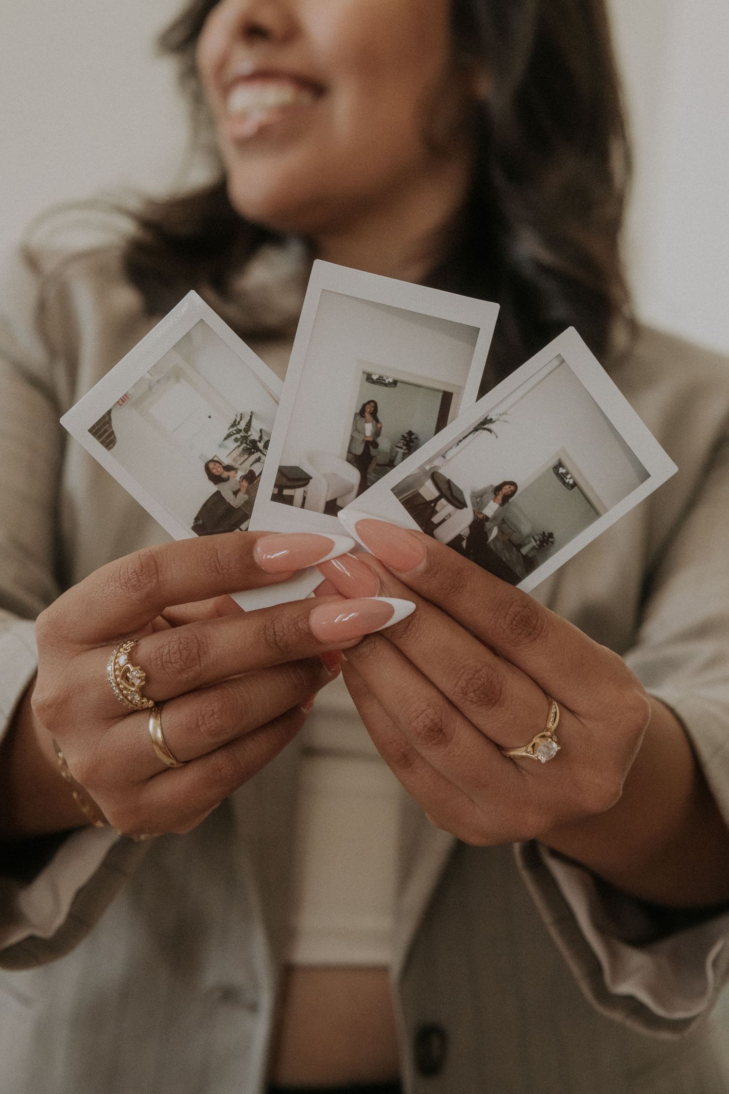 Smiling woman holding three Polaroid photos fanned out, wearing gold rings and French manicured nails, dressed in a beige blazer.