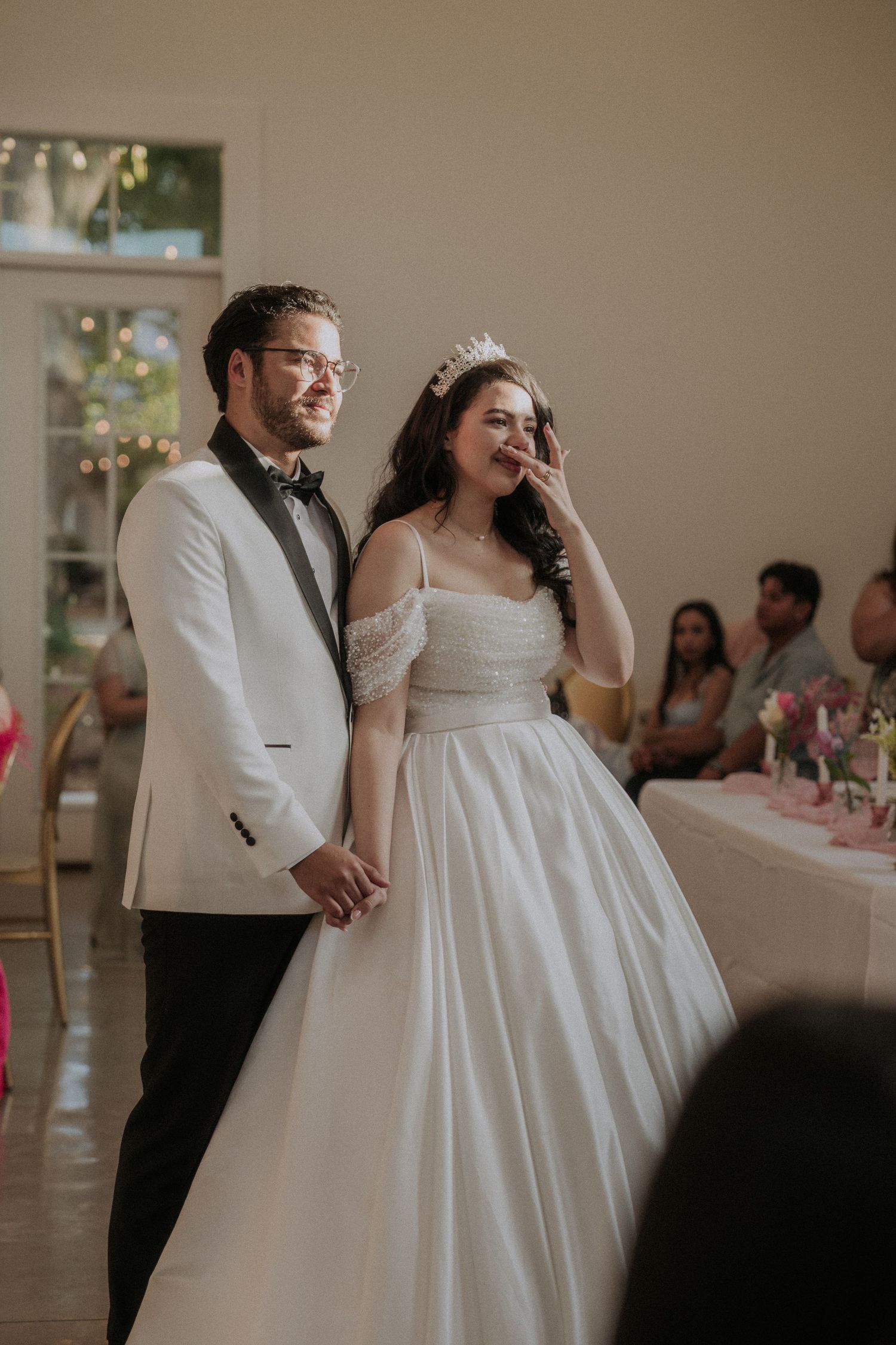 Smiling bride in tiara and groom in white tuxedo hold hands during wedding reception grand entrance.