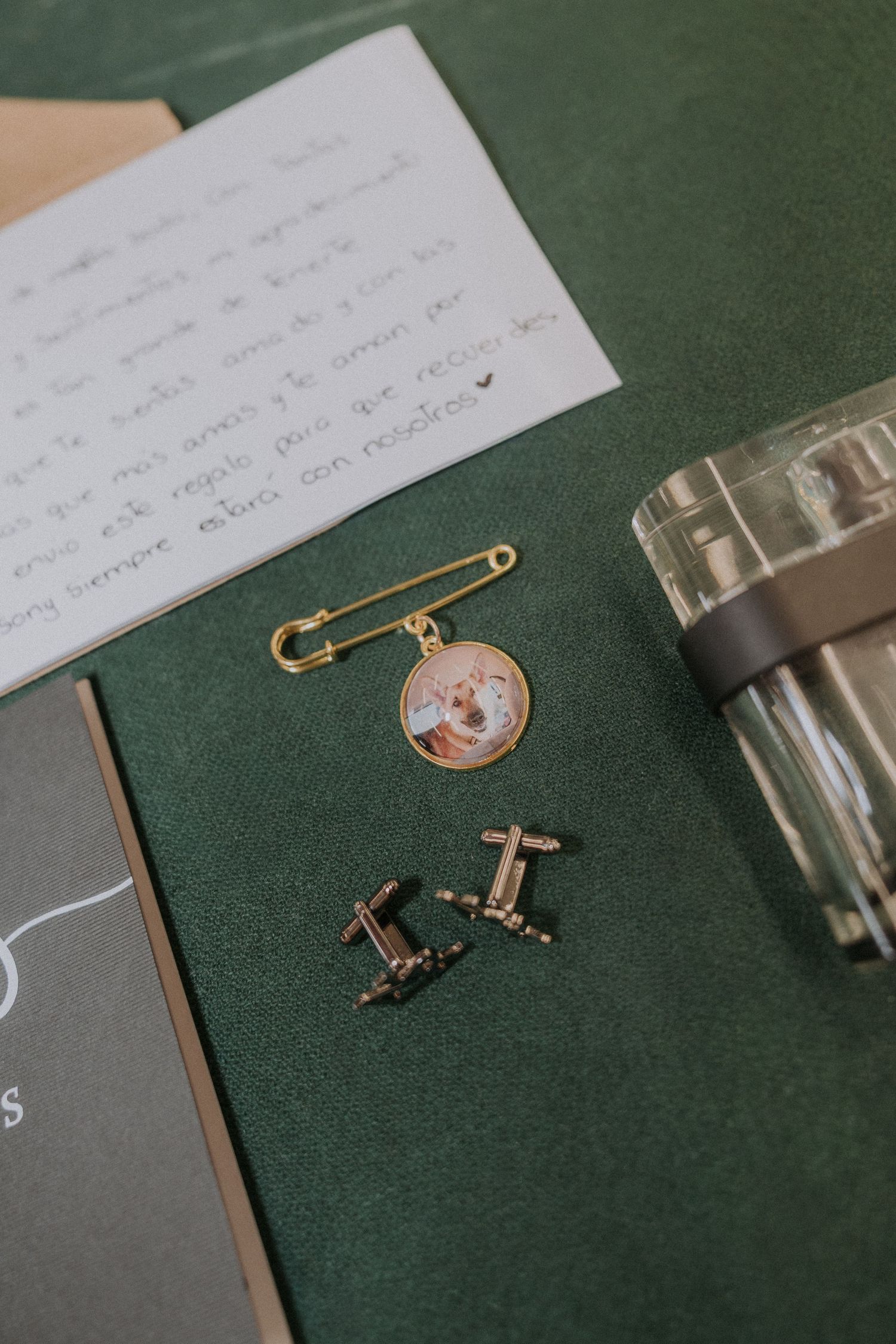 Gold safety pin brooch with dog photo charm, cufflinks, handwritten note on green velvet surface.