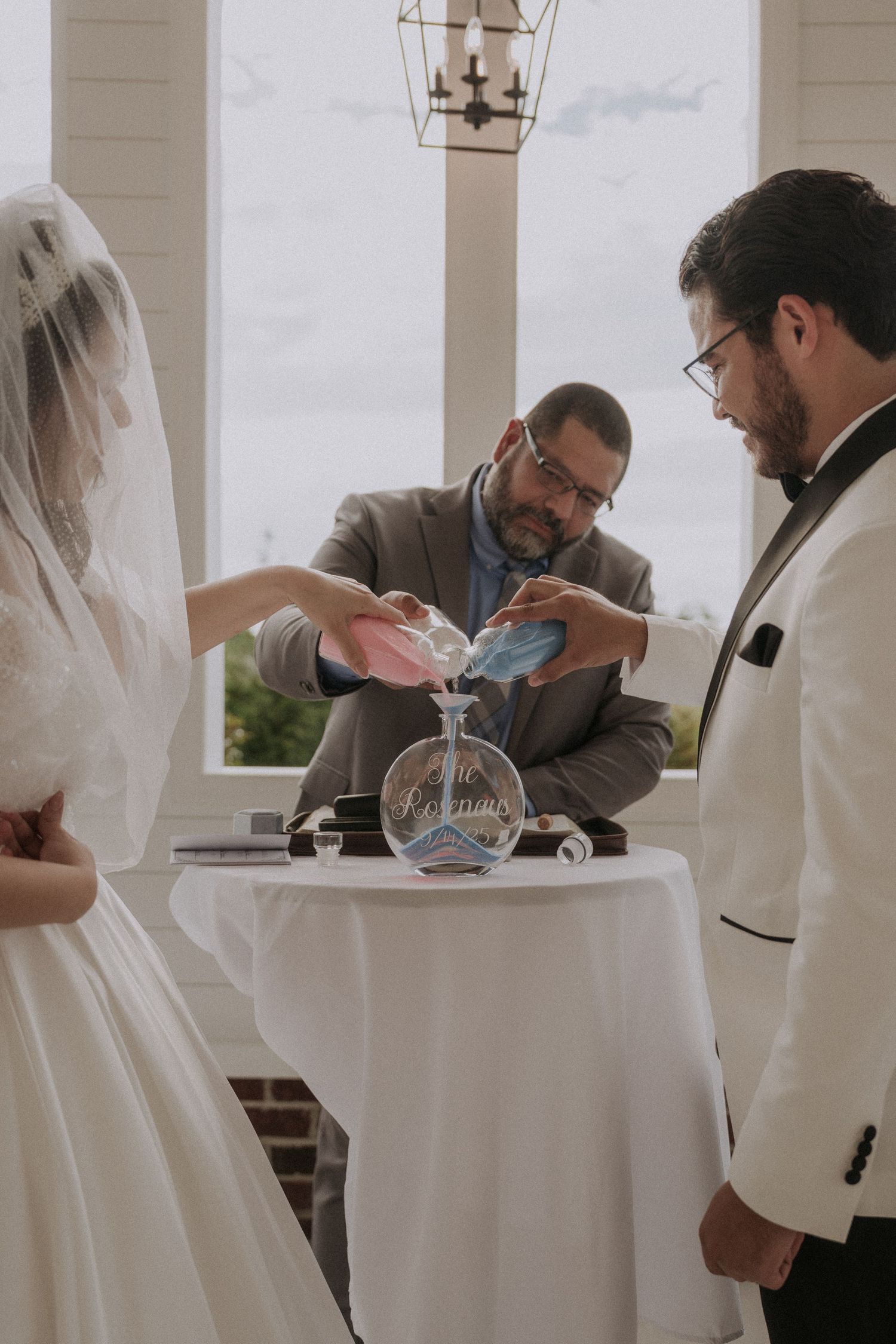 Bride and groom performing a unity sand ceremony at their wedding, officiated by a man in a gray suit.