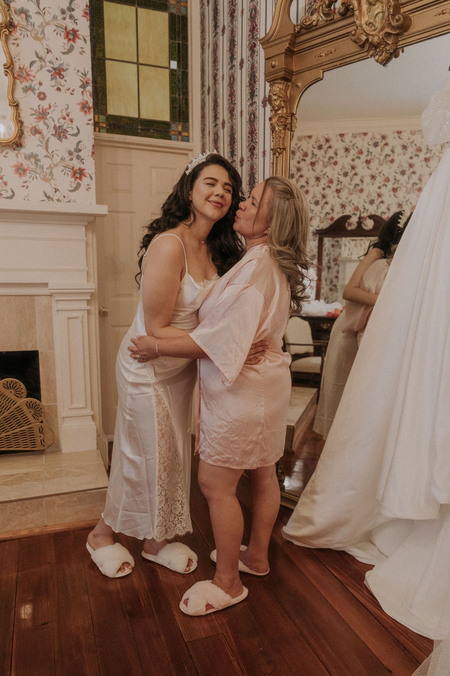 Bride in white slip dress hugging woman in pink robe during wedding preparations in elegant floral room.
