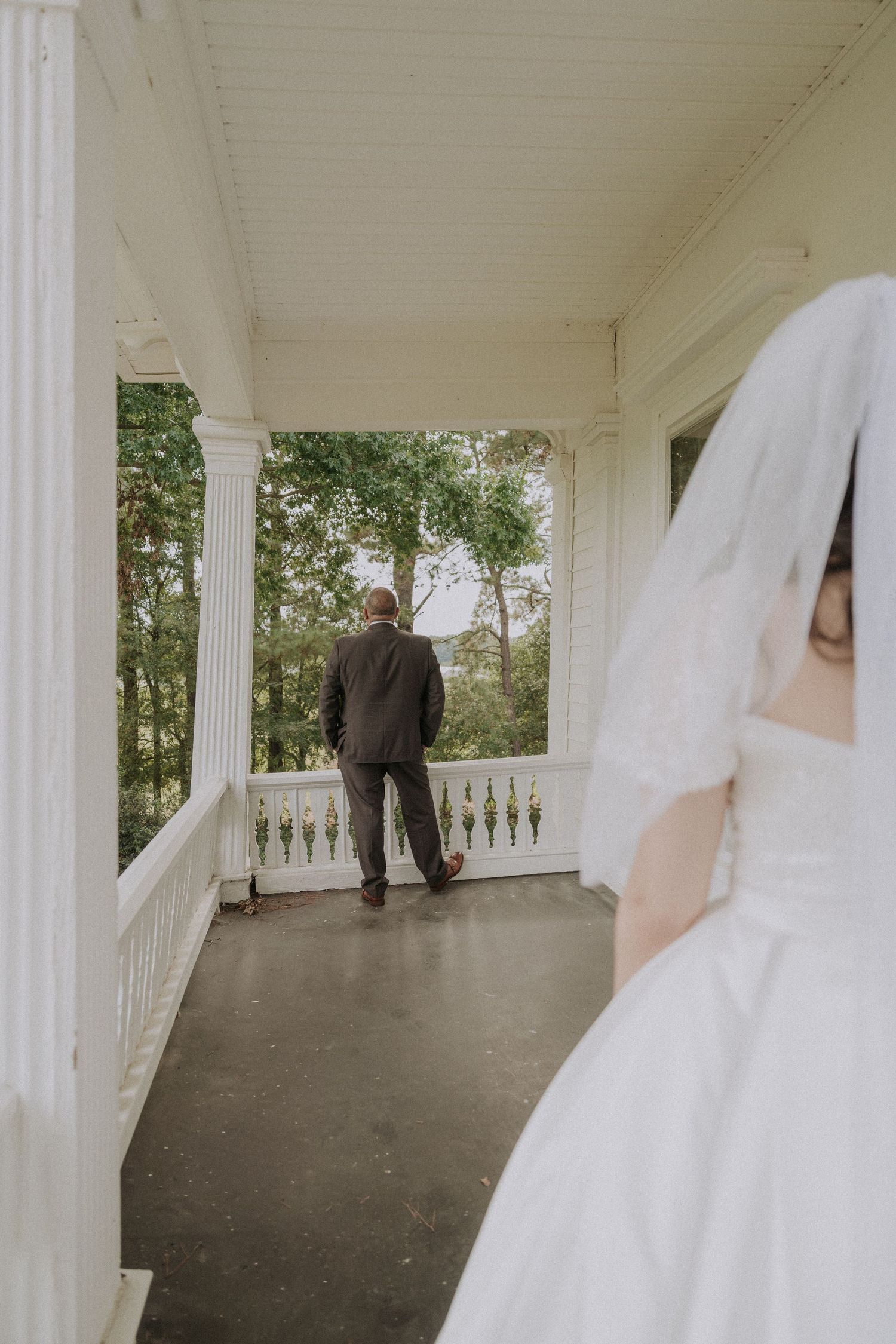 Bride in white gown and veil approaches groom standing on elegant white porch columns venue.