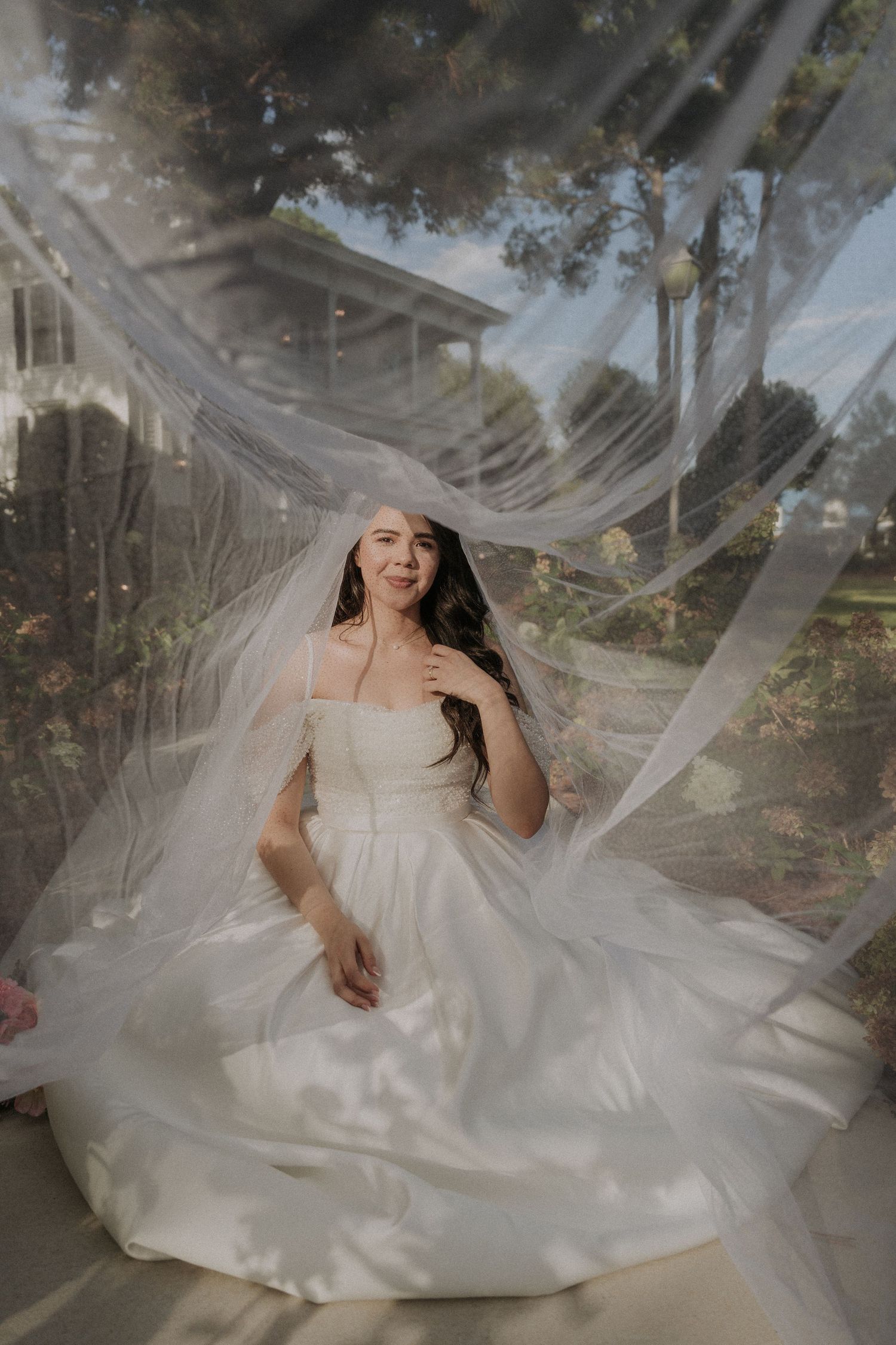 Bride in a white ball gown and veil seated outdoors surrounded by flowing tulle in a dreamy portrait.