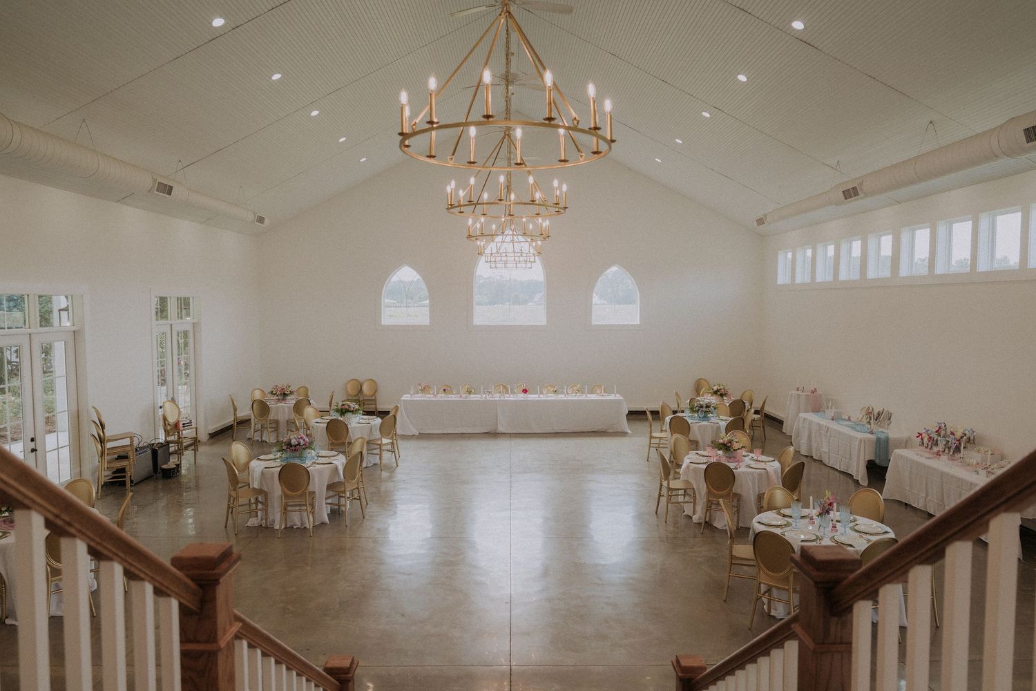 Elegant wedding reception hall with chandelier, round tables, and arched windows in a bright white venue.