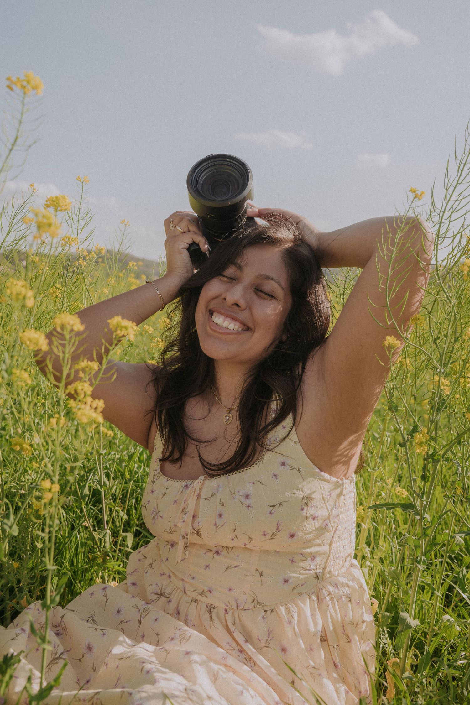 Smiling woman in floral dress sitting in yellow wildflower field holding camera above her head on sunny day.