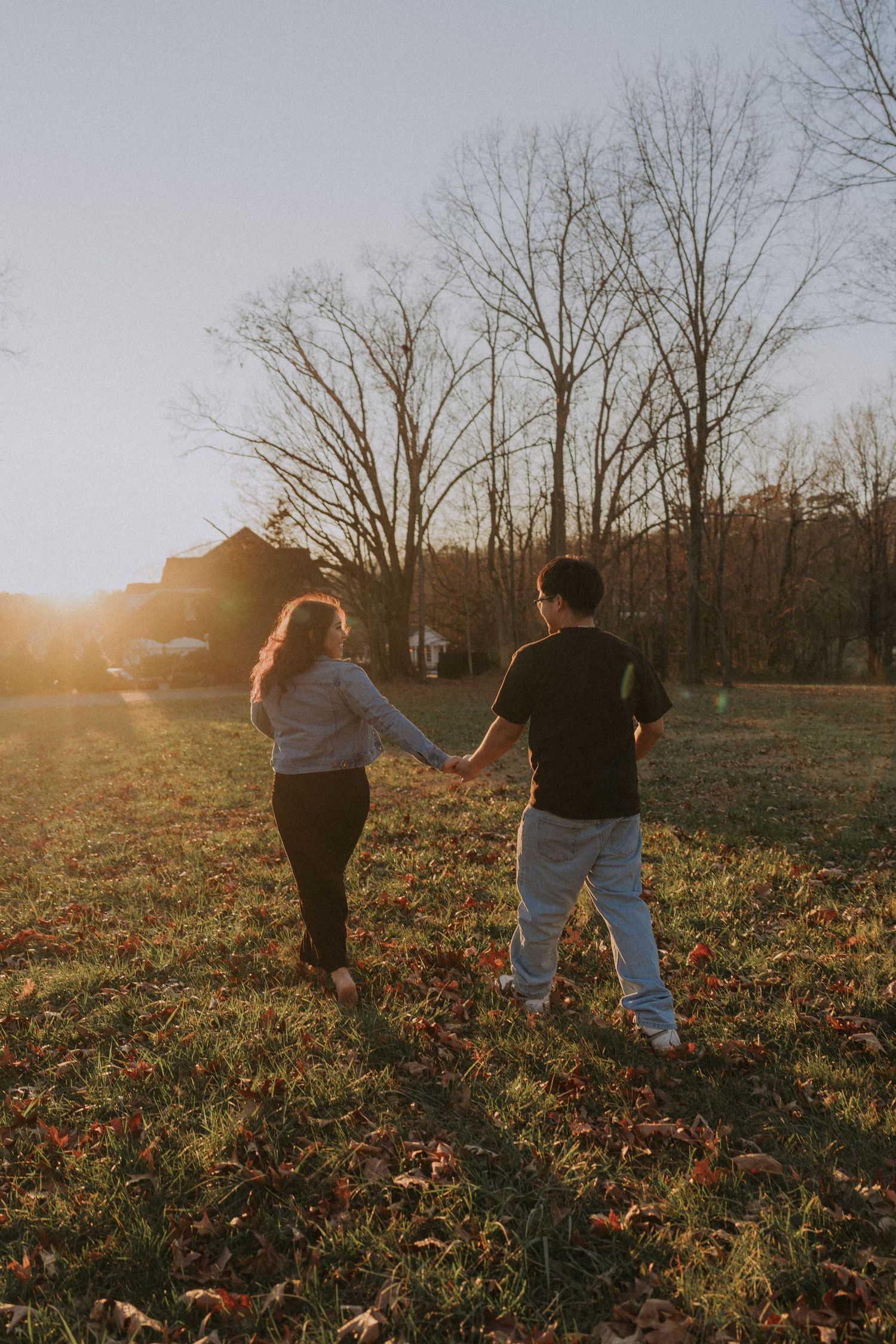 Couple holding hands walking through a sunlit autumn field with bare trees and golden hour light in the background.