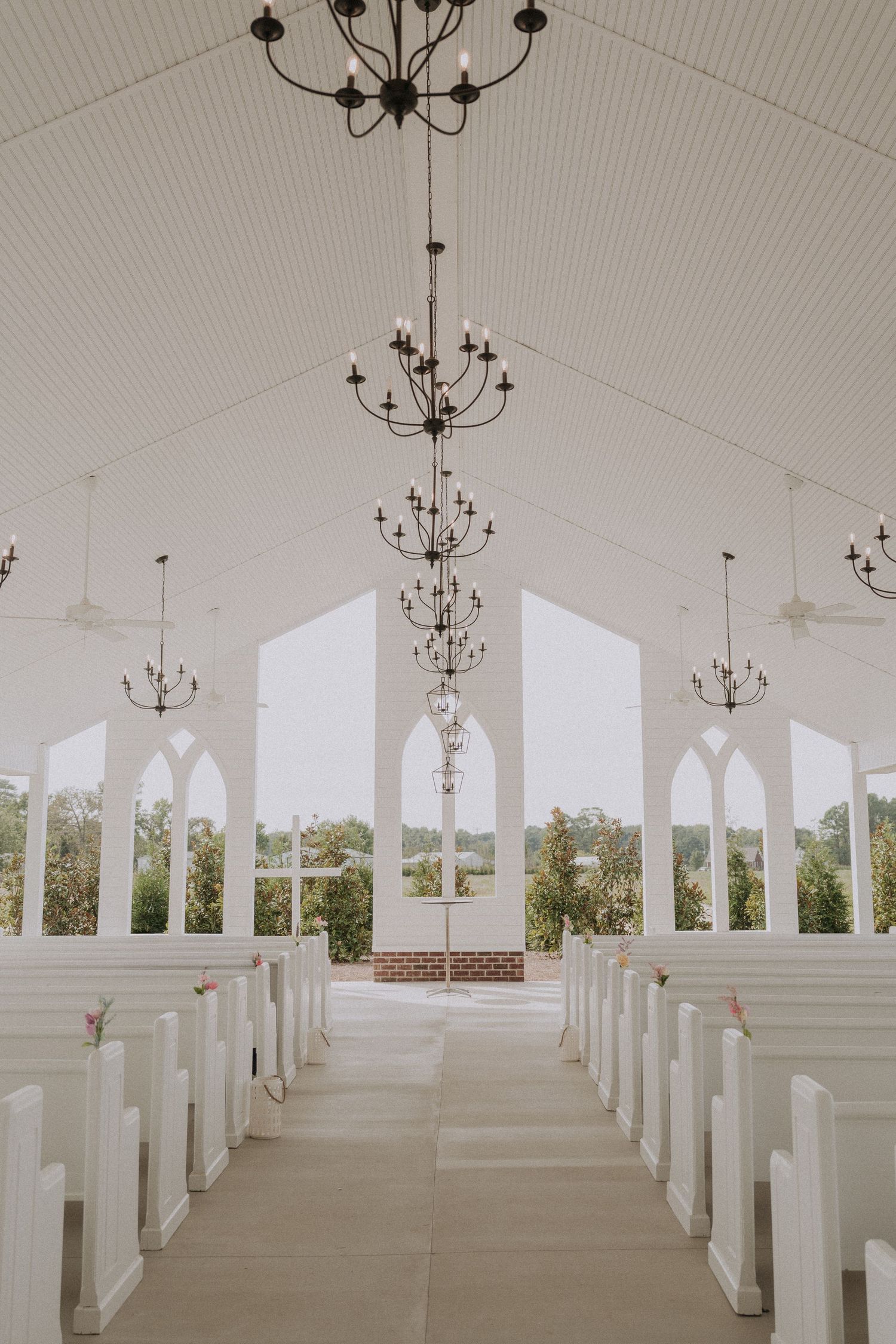 Elegant white chapel interior with chandeliers, arched windows, and wooden pews lining the aisle.