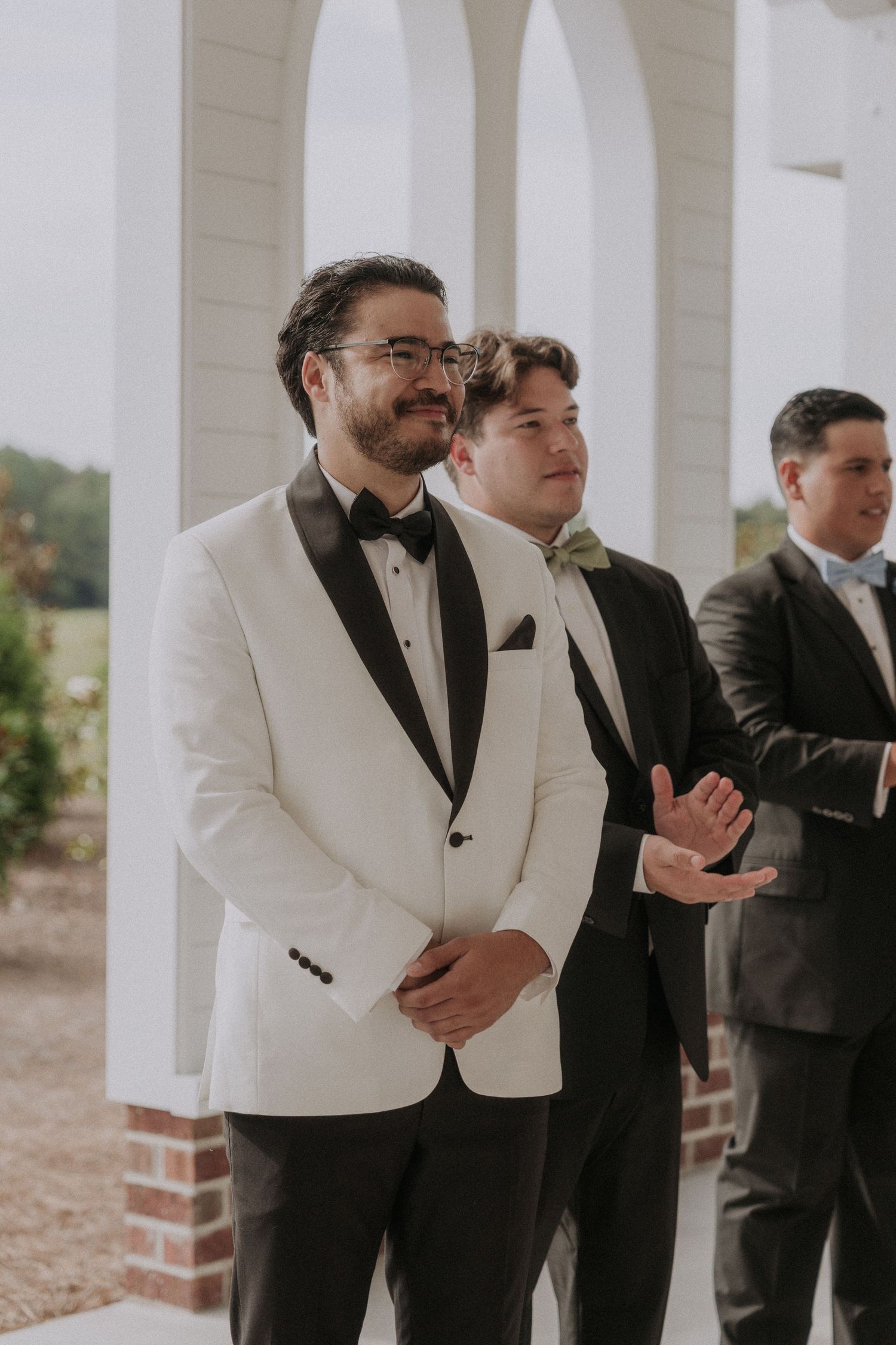 Groom in white tuxedo jacket smiling at altar with groomsmen in black suits behind him at outdoor wedding.