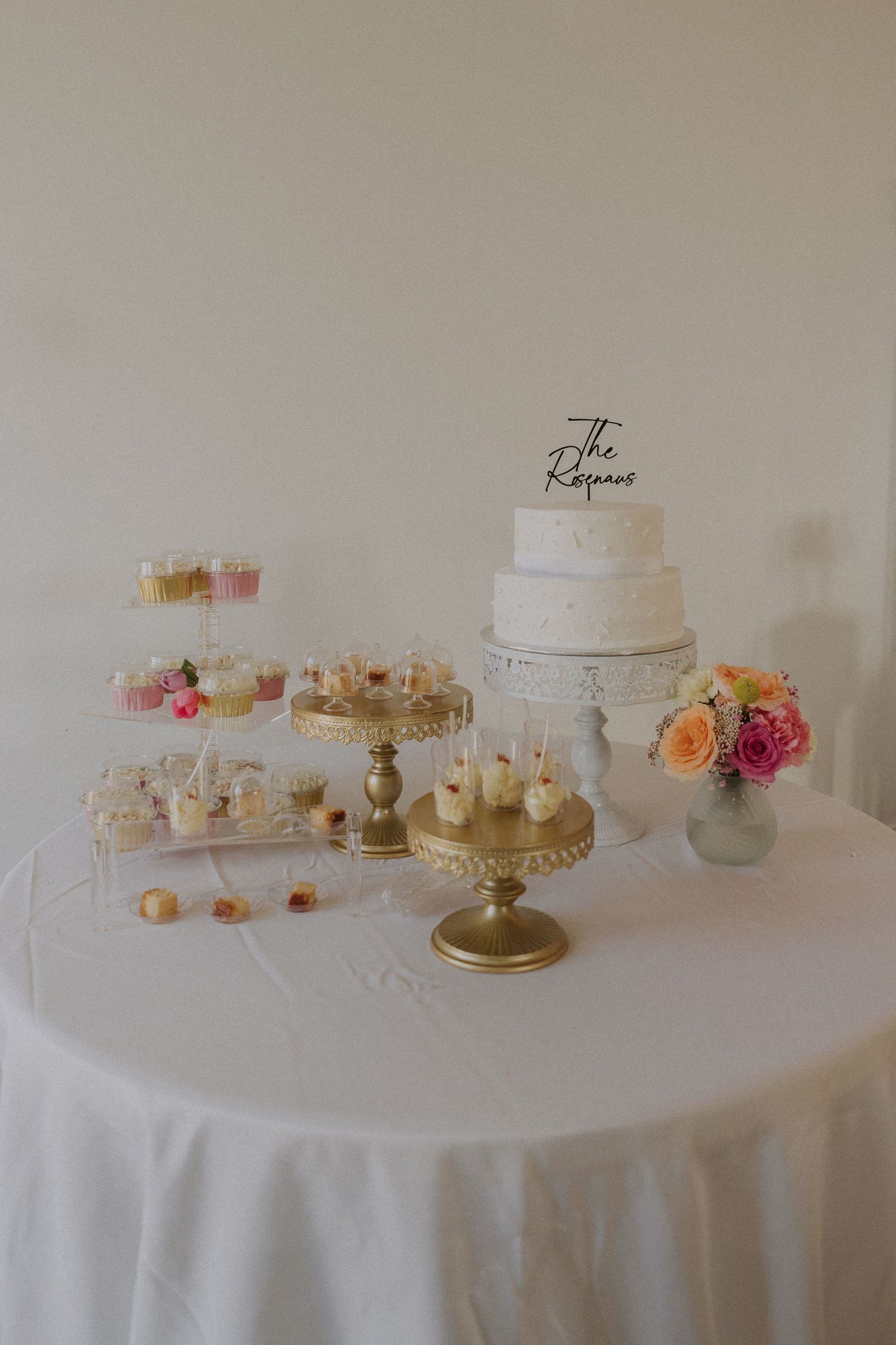 Elegant wedding dessert table with tiered white cake, gold cake stands, cupcakes, and fresh floral arrangement on white linen.