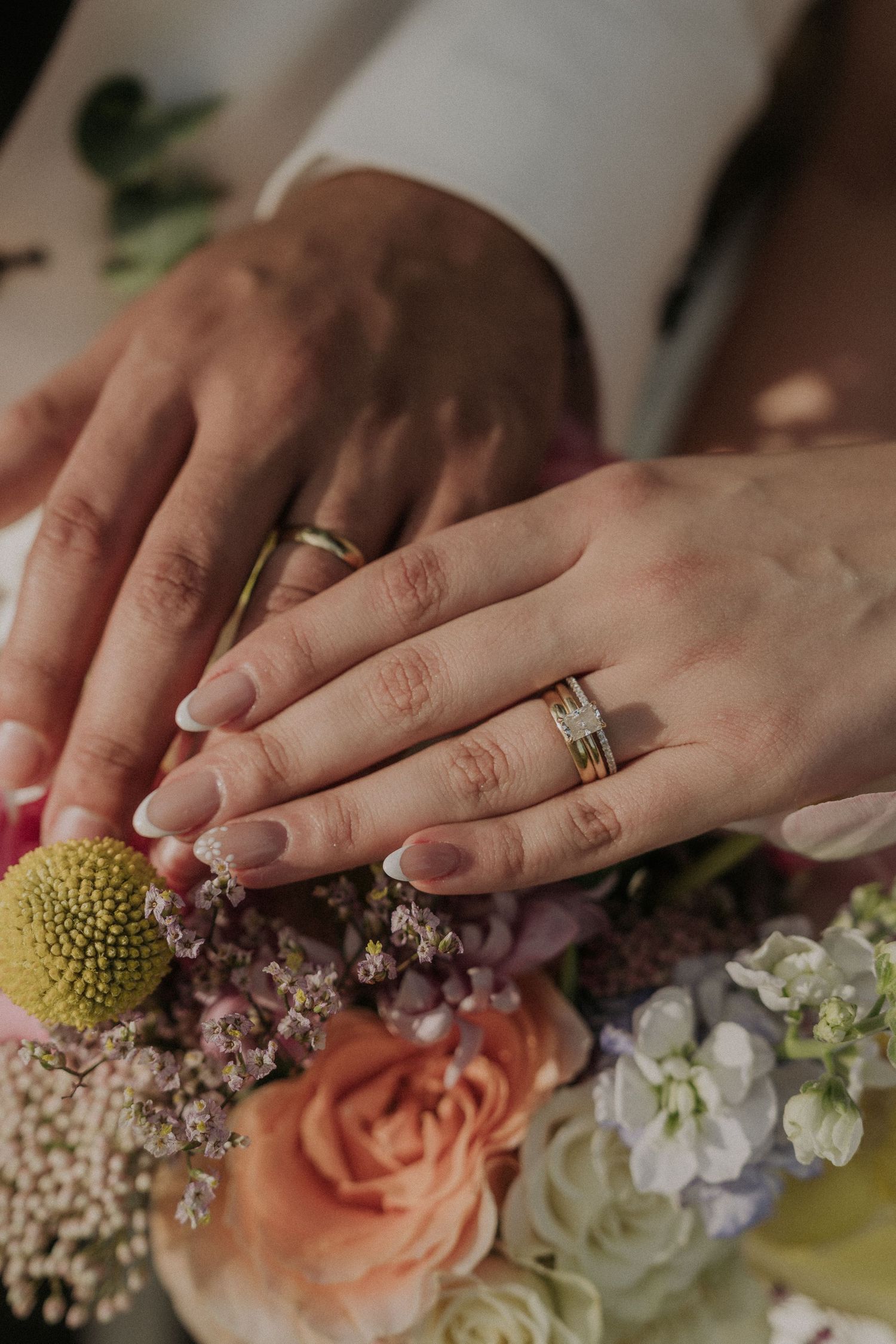 Close-up of newlyweds hands displaying wedding rings resting on a colorful bridal bouquet with roses.