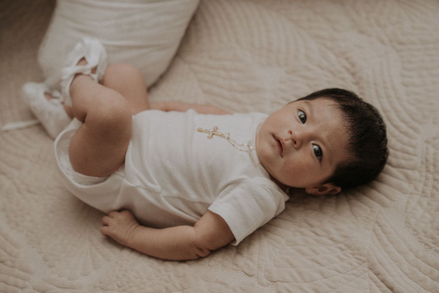 Baby wearing white clothing lies on cream colored bedding in a series of candid poses.