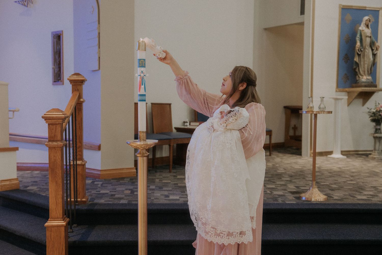 A woman in a pink dress holding a baby in a white christening gown while lighting a candle during a baptism ceremony.