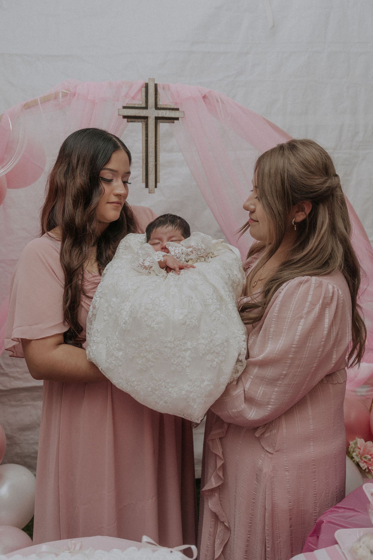 A baptism celebration with pink decor features a baby wrapped in white lace while being held near a wooden cross.