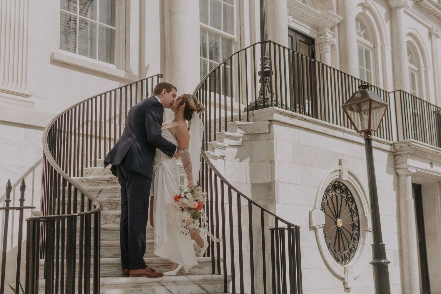 A couple poses on an elegant white staircase with ornate railings during their wedding.