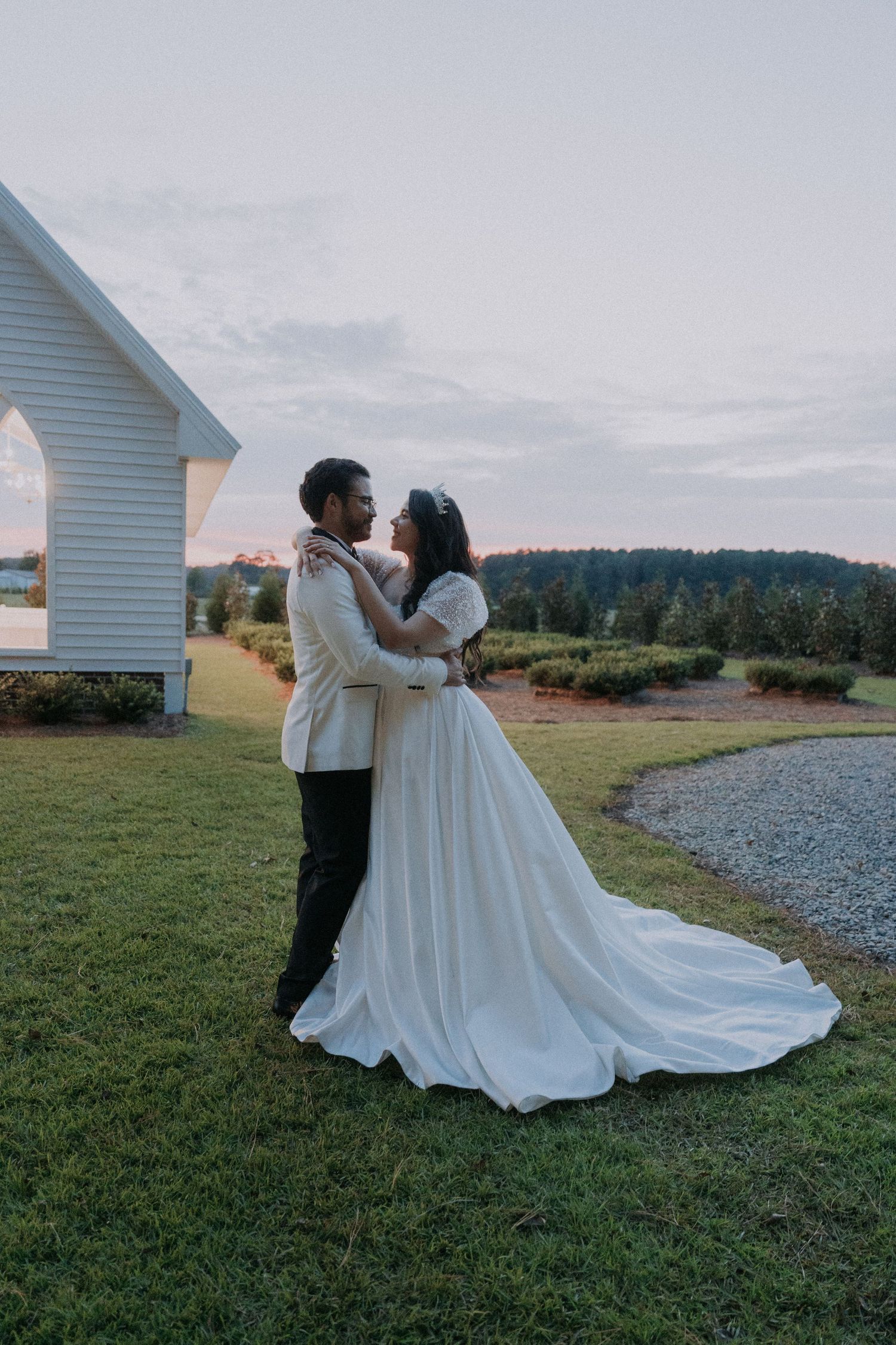 Bride and groom embrace at sunset near a white chapel, with rolling green hills in the background.