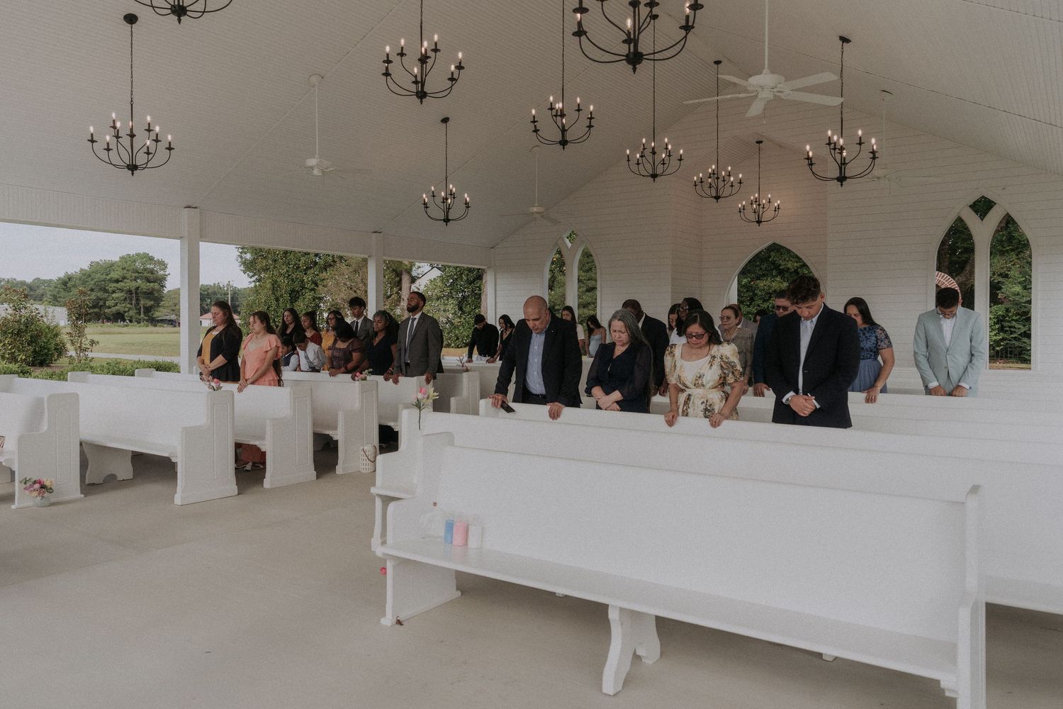 Guests stand in white pews inside an elegant open-air wedding chapel with chandeliers during a ceremony.