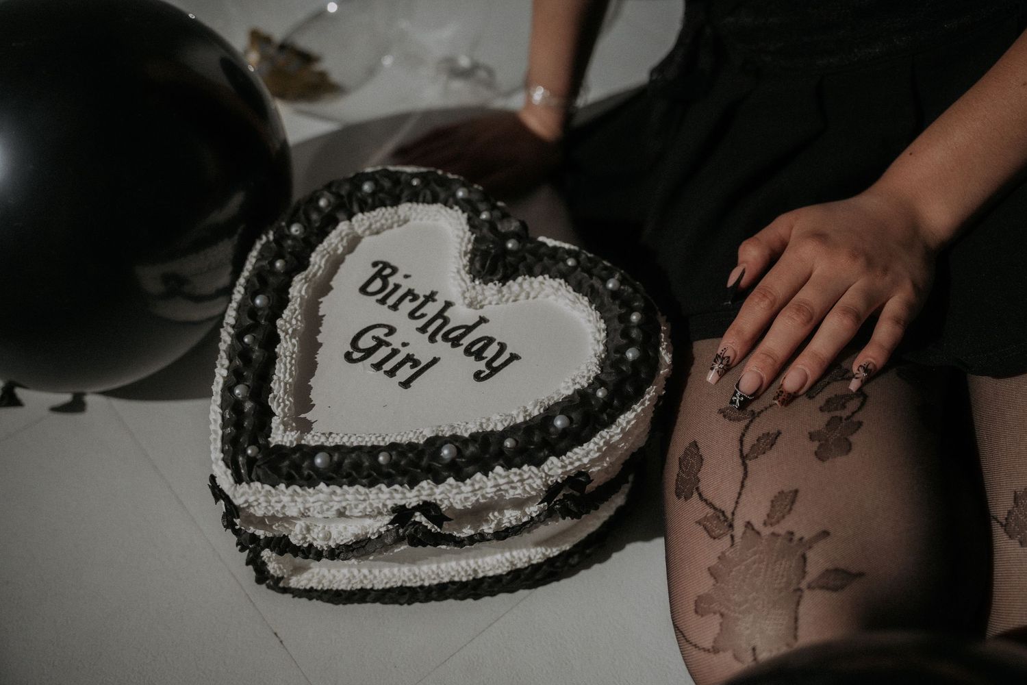 A heart-shaped birthday cake with dark frosting sits next to an arm on a white surface.