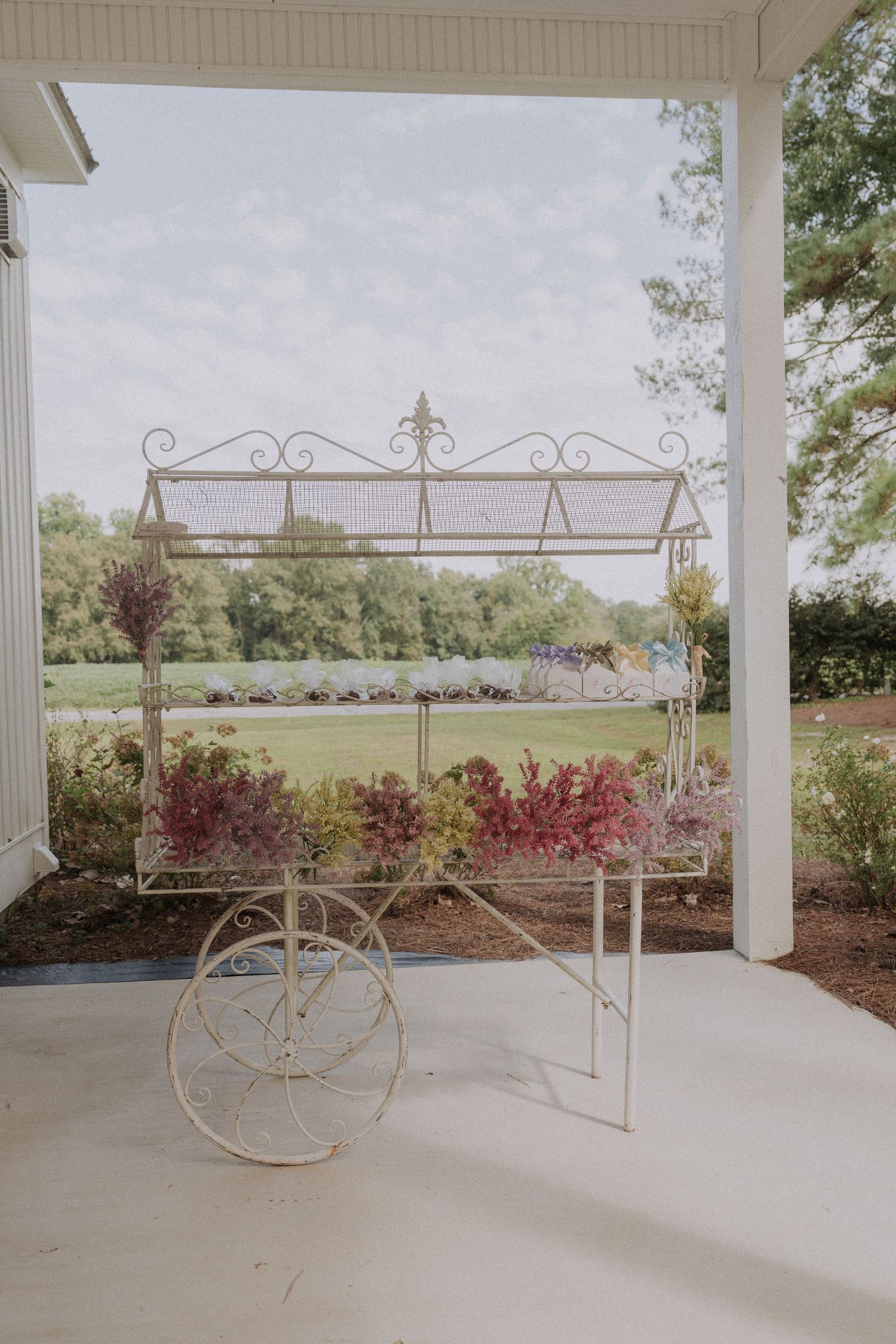 Vintage white ornate metal flower cart with colorful blooms on a porch overlooking a lush green garden.