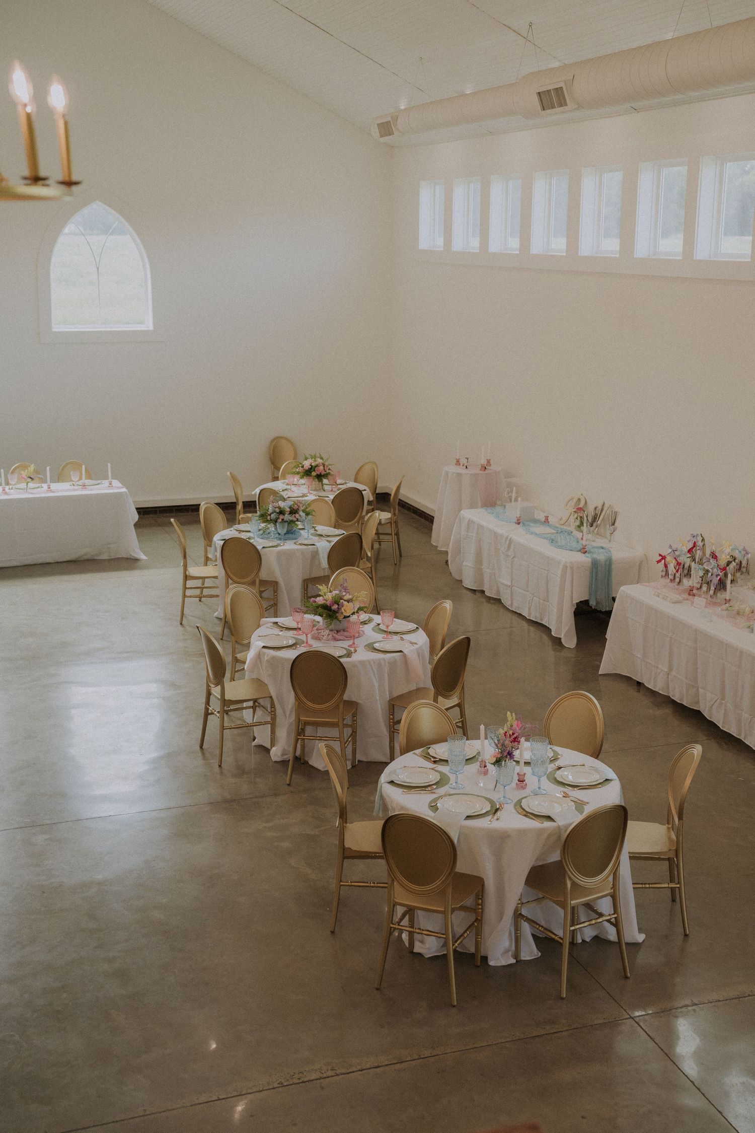 Elegant wedding reception hall with round tables, white linens, gold chairs, floral centerpieces, and soft natural lighting.