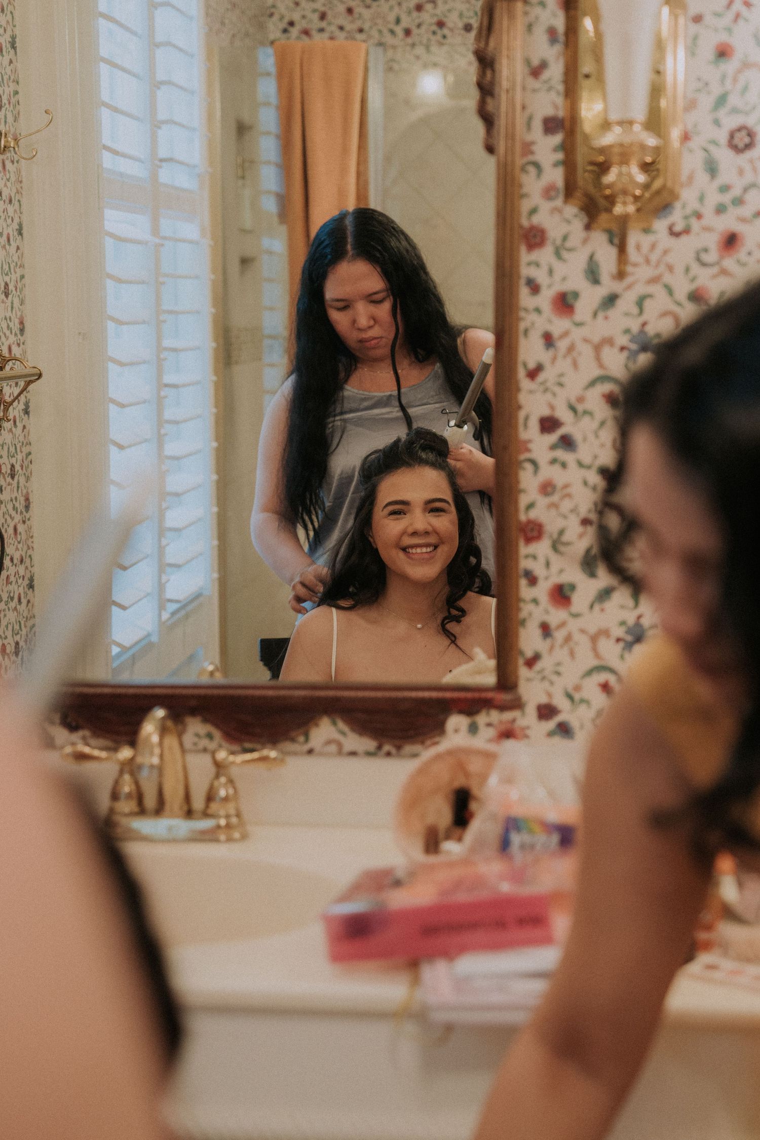 A woman smiling in a floral bathroom mirror while another styles her hair with a curling iron.