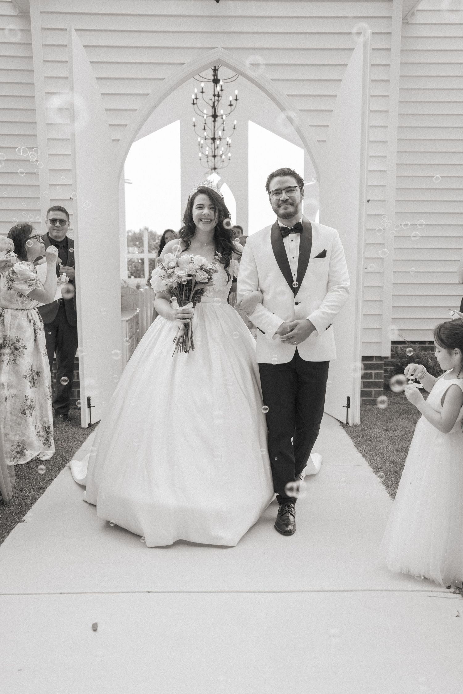 Bride and groom exit white chapel through arched doorway surrounded by bubbles in classic black and white wedding photo.