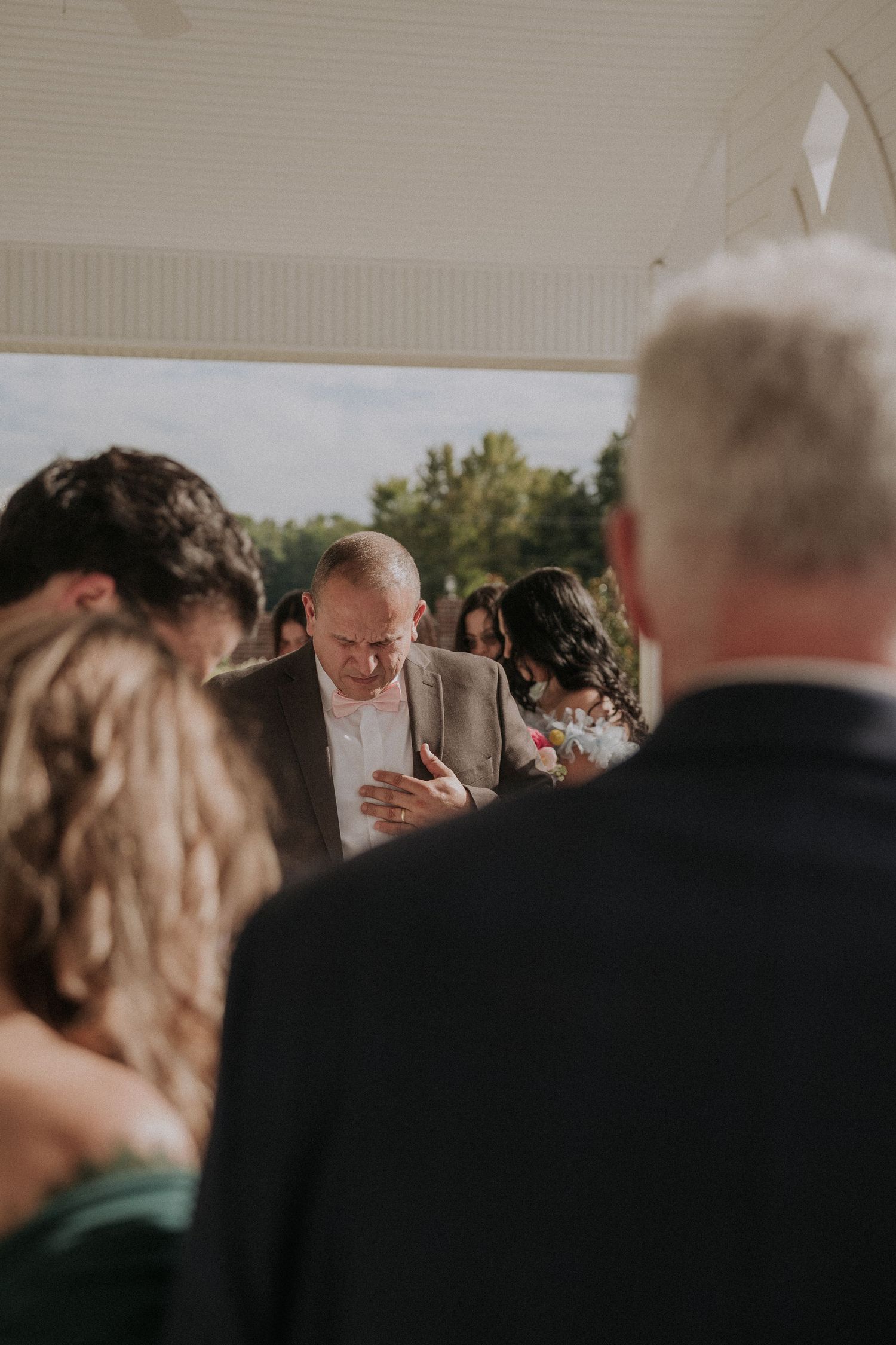 Wedding guests bow heads in prayer during outdoor ceremony under white pavilion.