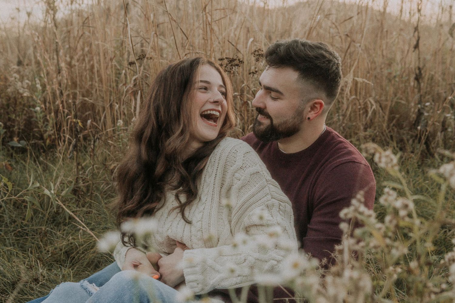 A couple shares intimate moments in a golden field during sunset, laughing and embracing in casual autumn attire.