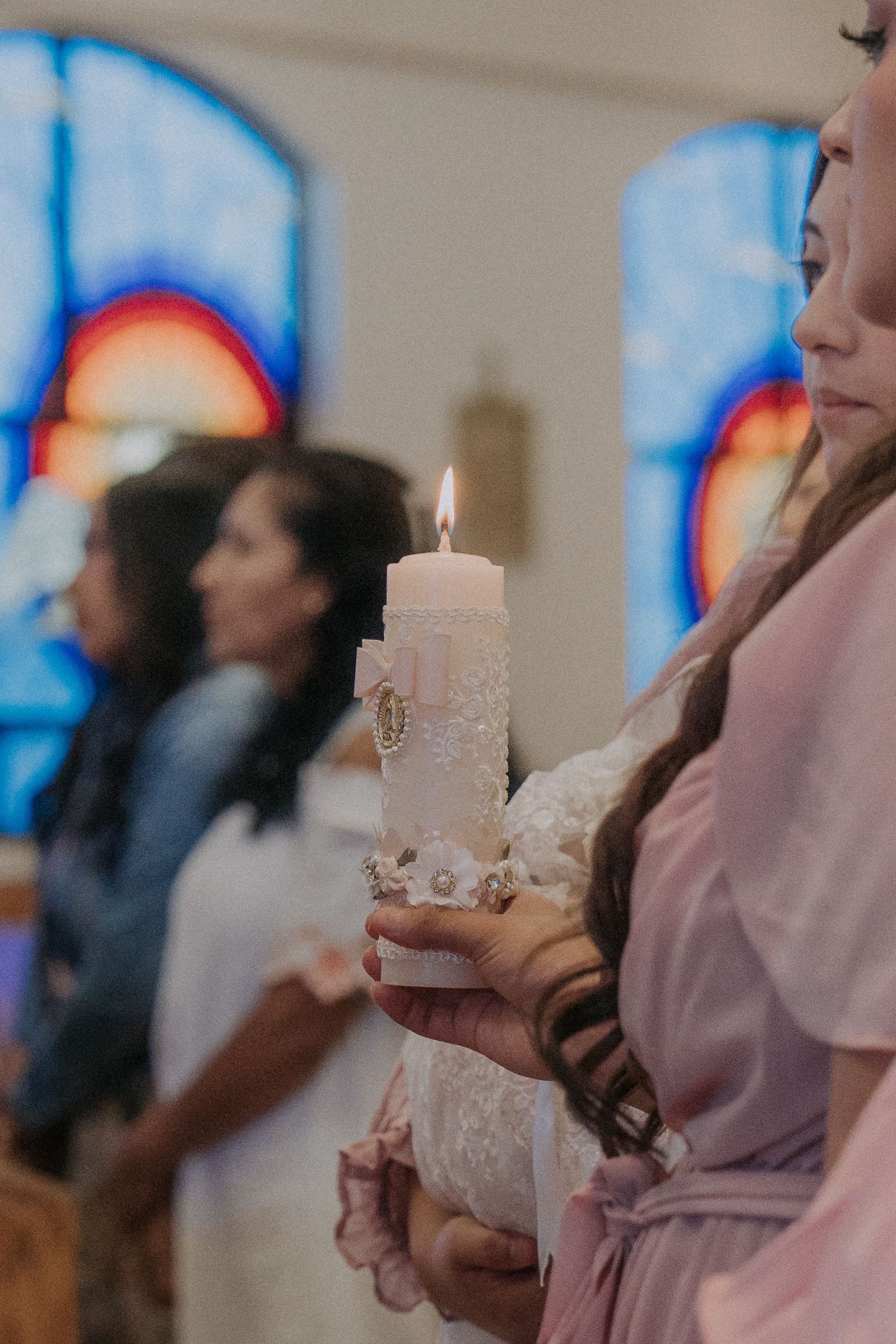 People holding decorated white candles during a baptism ceremony with stained glass windows in background.