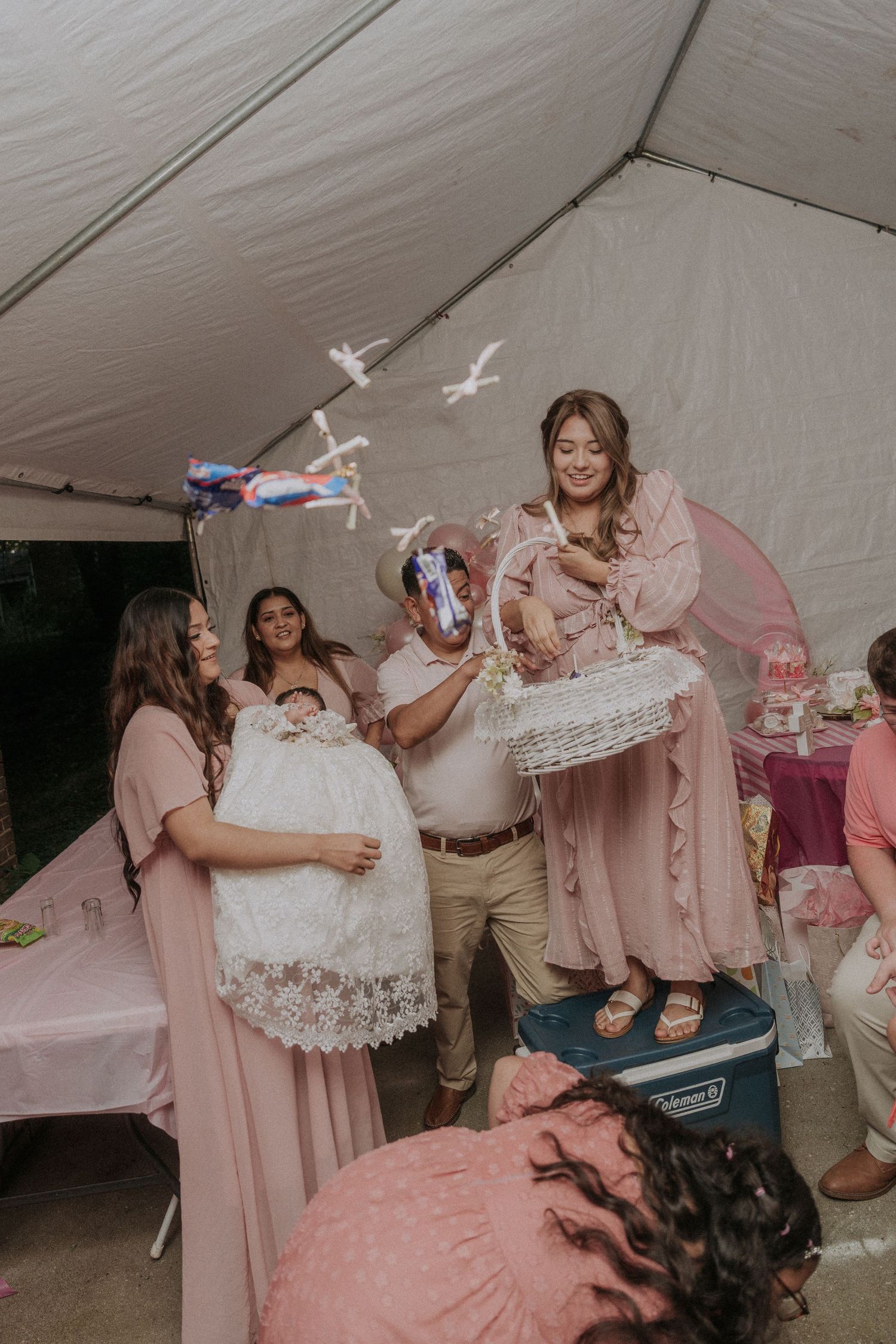 Party guests in pink attire gather around a cooler for an entertaining baptism reception game.