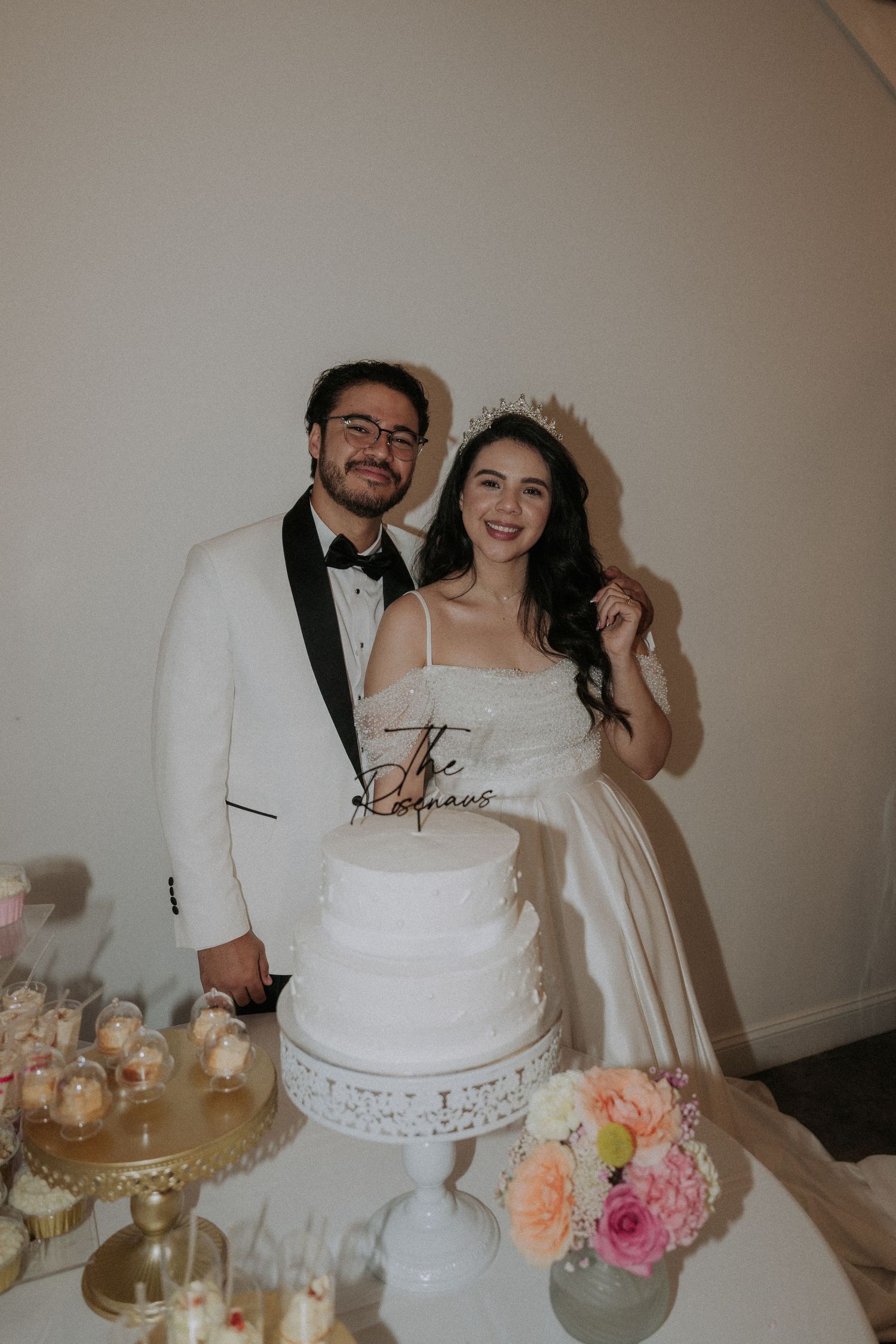 Bride and groom smiling beside a white three-tier wedding cake at their elegant reception table.