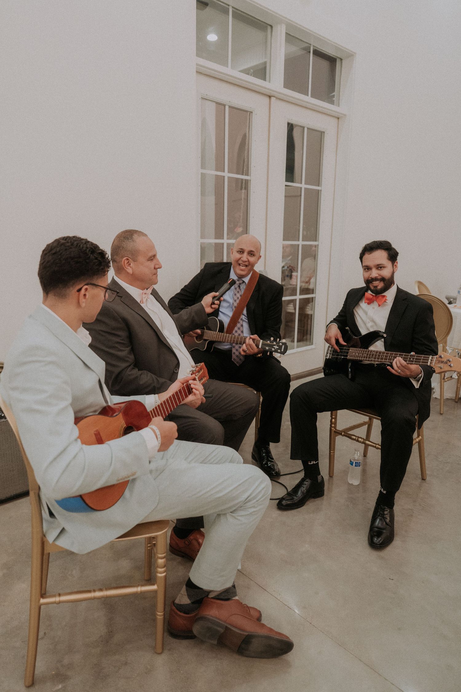 Four men playing guitars and singing together while seated in chairs at a wedding reception.